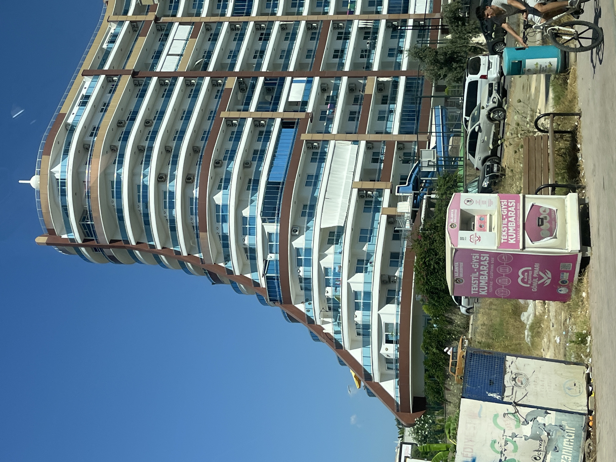 This image depicts a street scene in Alanya, Turkey, dominated by a distinctive, curved apartment building with multiple balconies and blue-tinted windows. In the foreground, a young man rides a bicycle past a pink textile donation bin. To the left, there are two dilapidated billboards with torn posters. The environment appears somewhat urban and well-lit under a clear blue sky.