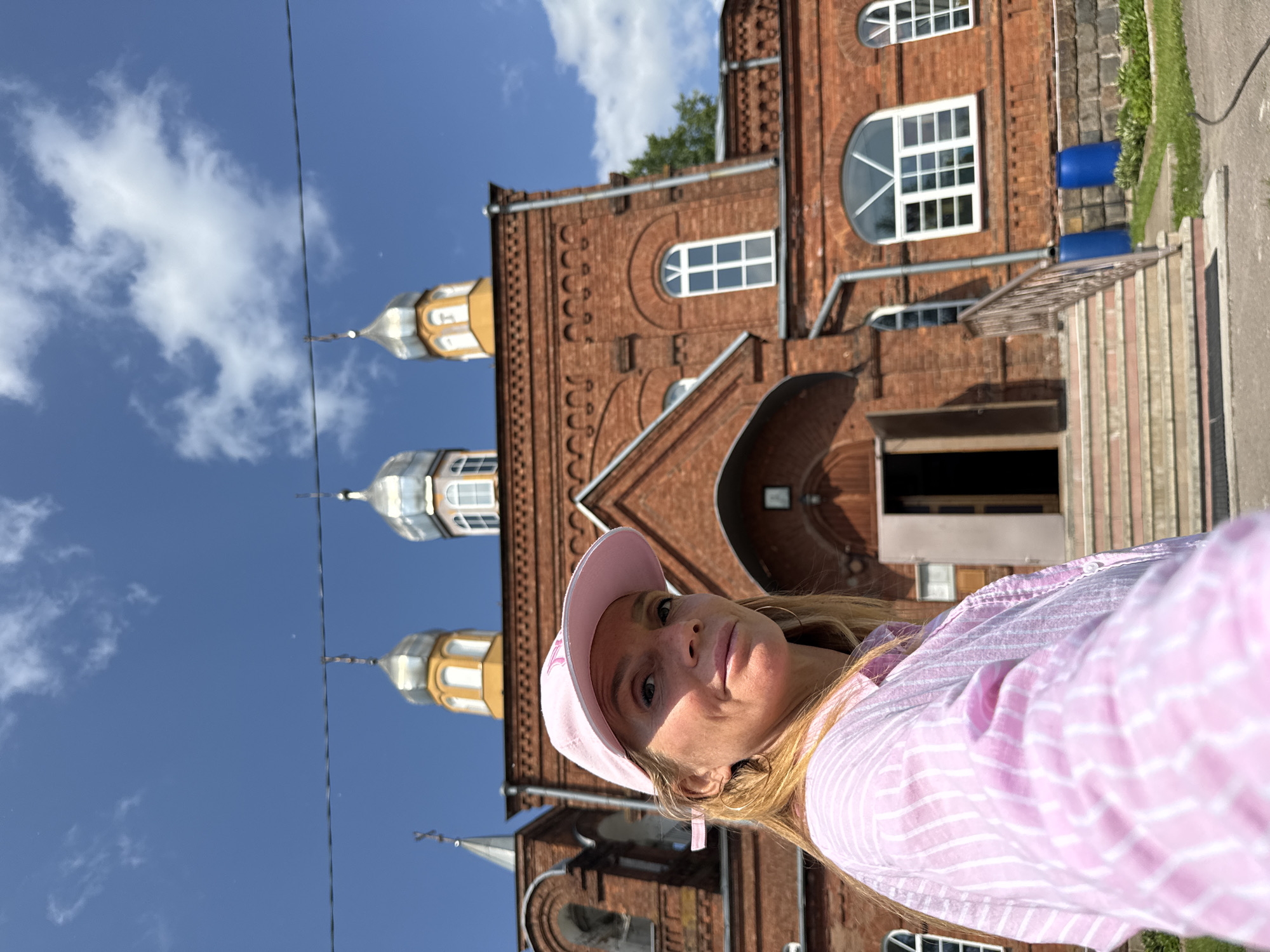 A blonde woman wearing a pink baseball cap and a pink and white striped shirt is taking a selfie outdoors. She is looking at the camera and smiling slightly. Behind her, a red brick building with a yellow-domed roof and multiple small windows is visible. The building appears to be a church or a religious institution. The sky above is blue with some scattered white clouds. The woman is positioned in the foreground, with the building in the background. The scene suggests a sunny day in Chudovo, Russia.