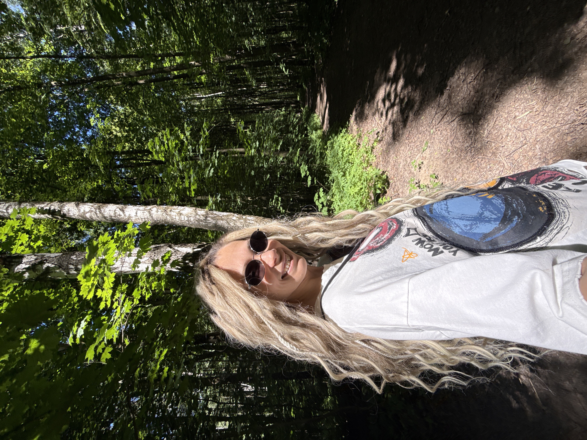 The image captures a female subject smiling in a sunlit forest in Rusanovo, Russia. The woman occupies the left side of the frame, positioned with her body slightly turned to the right and her face angled towards the camera, displaying a wide smile with visible teeth. She is wearing dark, oval-shaped sunglasses. Her hair is long, blonde, and predominantly wavy, interspersed with several lighter-colored sections that appear to be dreadlocks or braids. She wears a light-colored, long-sleeved t-shirt adorned with a large, multi-colored graphic print on the chest, featuring a prominent blue circular element and other abstract designs. A dark strap, possibly from a bag, is visible over her right shoulder.
The background is a dense forest characterized by numerous tall trees with abundant green foliage. Sunlight filters through the canopy, illuminating parts of the leaves and creating distinct patterns of light and shadow on the forest floor. A winding dirt path or trail extends into the distance through the trees on the right side of the image. The ground is covered with earth, scattered dry leaves, and patches of green undergrowth, particularly noticeable on the left side of the path. The trunks of several trees, including some with lighter bark, are visible throughout the wooded area.