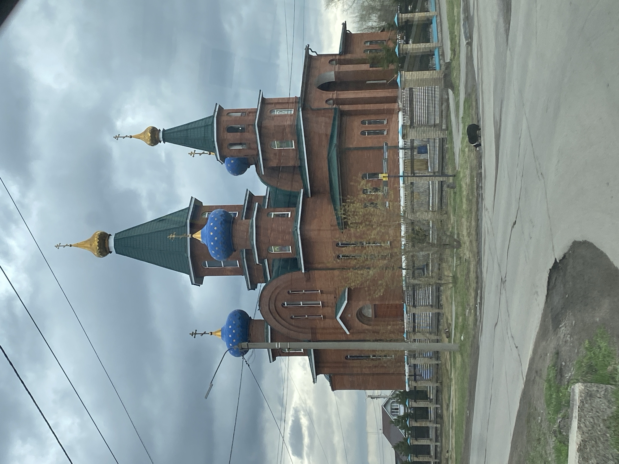 The image depicts a Russian Orthodox church located in Usolye-Sibirskoye, Russia. The church is constructed primarily of red brick with distinctive green roofs and onion-shaped blue domes adorned with gold stars. The building features multiple levels and a variety of architectural details, including arched windows, decorative moldings, and crosses atop the domes. In the foreground, there is a paved road with cracks and patches. Utility poles with overhead wires are visible on the left side of the image. The sky is overcast with grey clouds. To the side of the church there are fences and greenery, suggesting the proximity of a residential area. A dog is observed in the middle of the road.