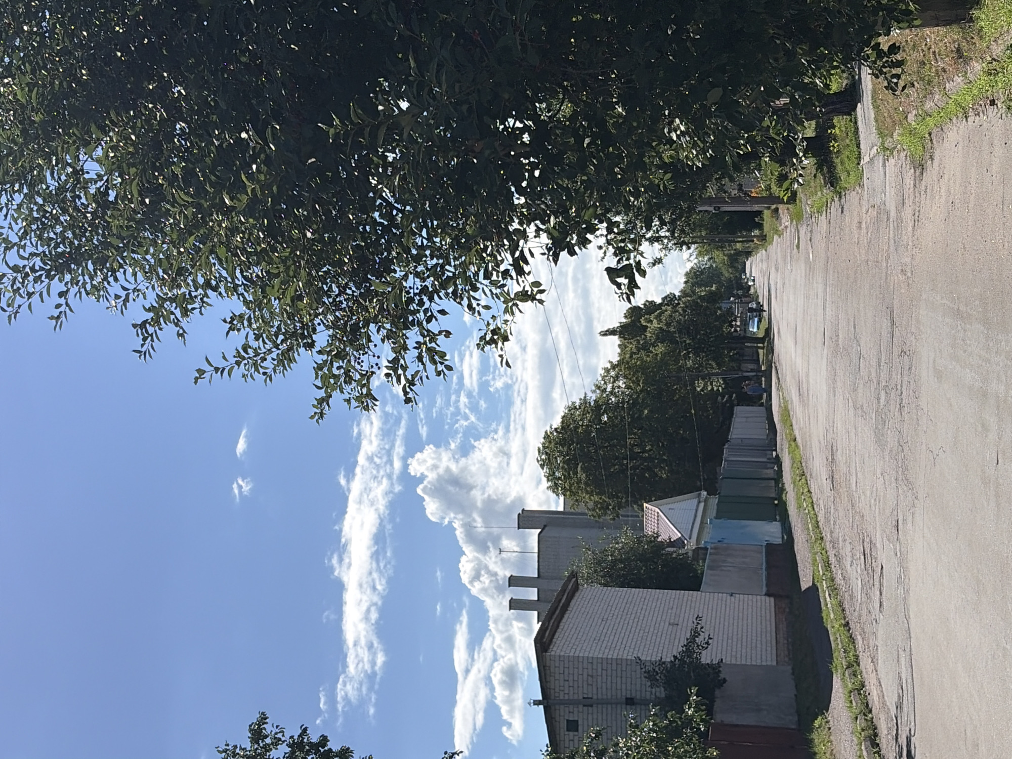 The image captures a street scene in Chernihiv, Ukraine, on a sunny day with a blue sky and scattered white clouds. The perspective is from the middle of a paved, somewhat cracked road. On the left, a white brick building with a grey foundation and a tall chimney is visible. Adjacent to it is a fence composed of various colored sections, including blue, green, and grey, with a small white house with a red-tiled roof behind it. Trees line both sides of the street, with dense green foliage dominating the right side of the frame. In the distance, several other trees and buildings can be seen. A few cars are parked or moving on the road, and a person can be seen walking in the far distance on the right. The environment appears to be a residential or semi-industrial area within the city.