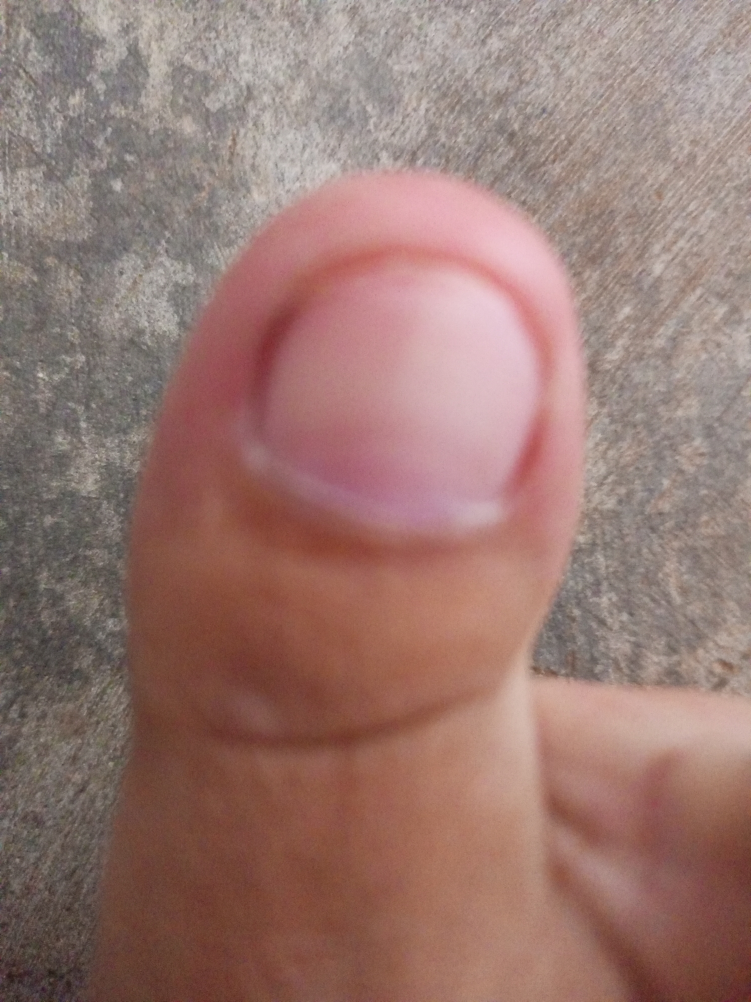 The image is a close-up, blurry shot of a thumb against a textured, gray surface. The thumb is pale pink with a visible nail. The gray surface appears to be concrete or asphalt with a rough texture and some lighter speckles. There is no discernible indication of the location being in Batam, Indonesia, or any urban environment. The scene is devoid of any human figures other than the partially visible thumb and hand. No actions or interactions are depicted.