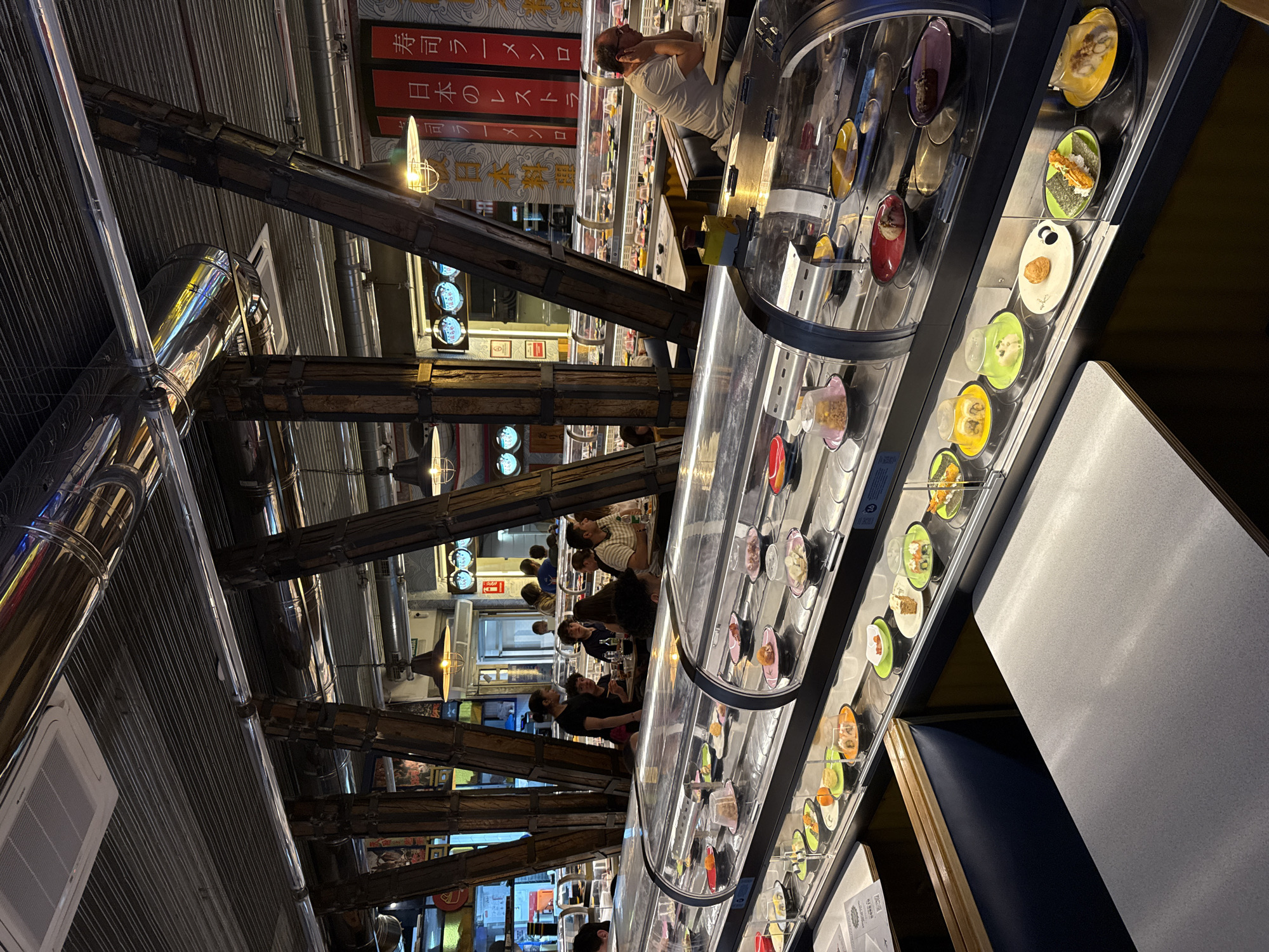 The image shows the interior of a Kura Revolving Sushi Bar in Madrid, Spain. The restaurant features a conveyor belt system carrying plates of sushi. Customers are seated at tables along the conveyor belt. Structural wooden beams and metal pipes are visible on the ceiling. Patrons are seated, eating sushi. Signs with Japanese writing are visible in the background.