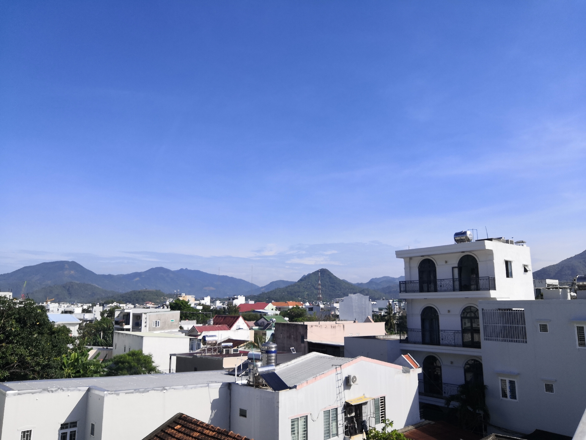 This is a daytime, eye-level, medium-shot photograph of a residential area in Bac Nha Trang Ward, Vietnam. The scene is dominated by a clear, bright blue sky. In the foreground, several white buildings with flat roofs and varying architectural styles are visible. Some buildings feature multiple stories, balconies with decorative railings, and arched windows. One building on the right has a prominent solar water heater on its roof. Various trees with dark green foliage are interspersed between the buildings. In the background, a range of lush green mountains stretches across the horizon, partially obscured by a haze. The overall impression is of a sunny day in a densely populated urban area with a mountainous backdrop.