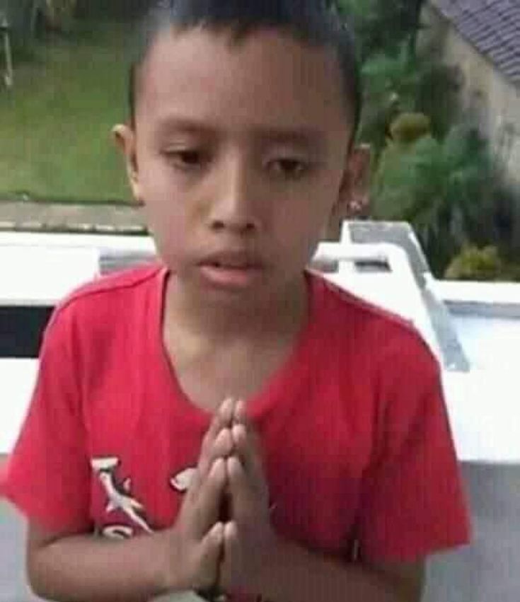 The image shows a young boy in a red t-shirt with a graphic on it. He is praying with his hands clasped in front of his chest. He is looking down at his hands. His face is serious. The image is likely taken in Geneng, Indonesia, based on the provided coordinates (-7.45472, 111.427584). The background shows a partially visible white wall and some green foliage. The boy's environment appears to be a residential area.