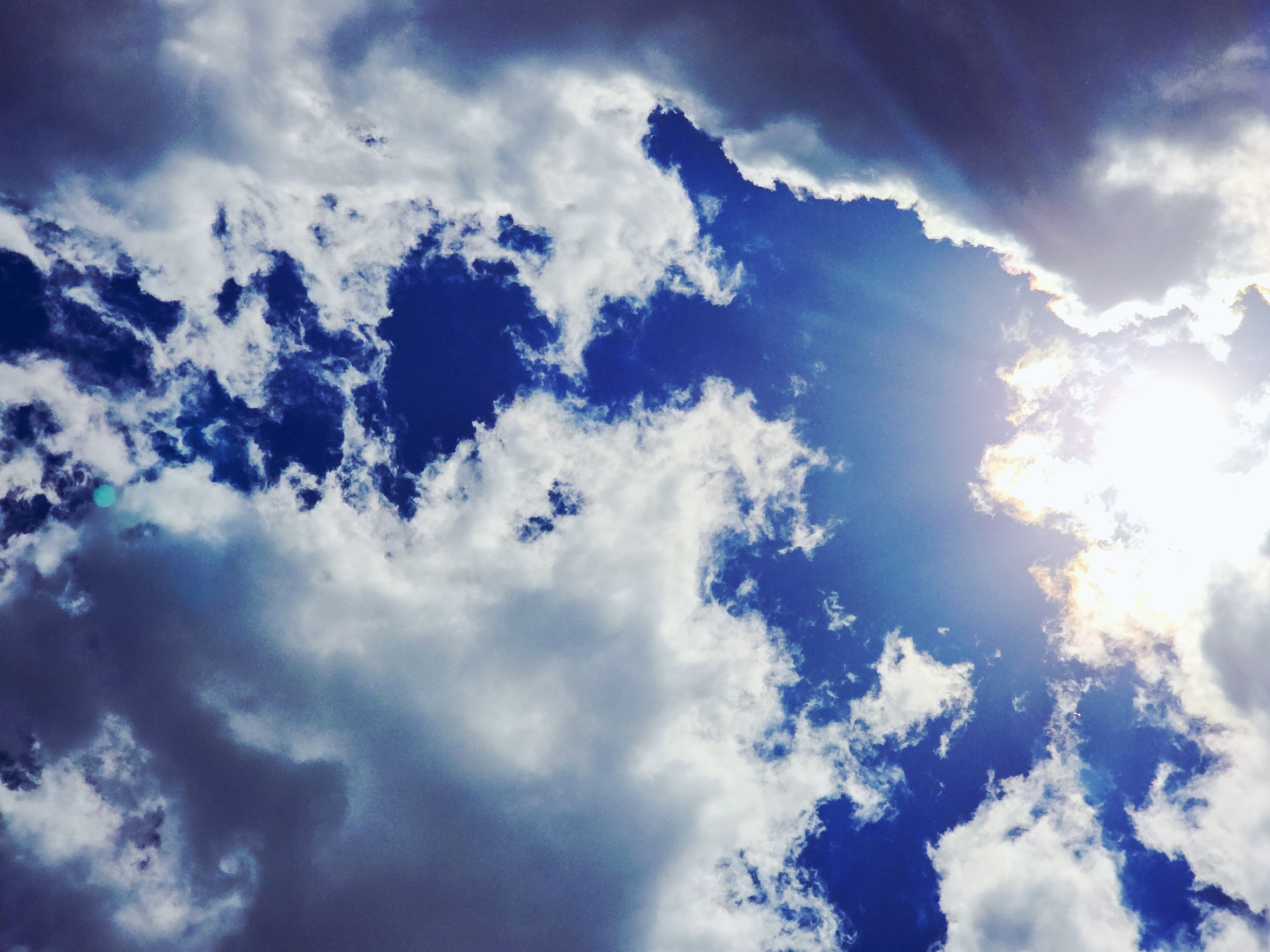 The image depicts a sky scene in an unknown city with blue patches amidst white and gray clouds. Sunlight filters through the clouds on the right side, creating a bright glare. The sky ranges from a deep blue to lighter blue shades.