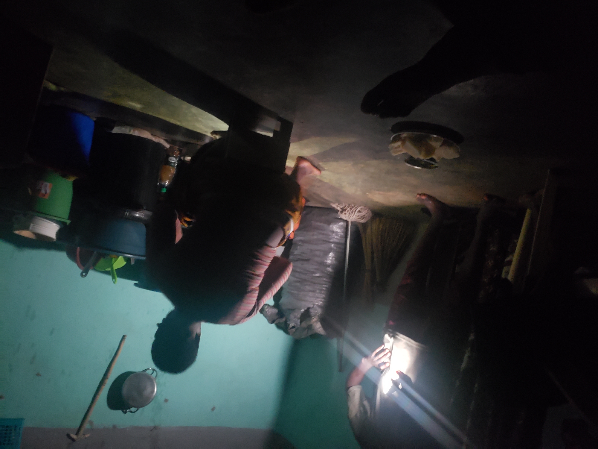 The image depicts an indoor scene, likely a room, with poor lighting conditions. The walls are painted a light blue color. In the foreground, a person is crouched down, facing away from the camera. They are wearing a striped shirt. To the left, another person is standing and partially visible, illuminated by a bright light source. This person is wearing a light-colored shirt and darker lower garments.
On the floor, a plate with some food items, possibly bread or yams, is visible. A broom made of natural fibers leans against a large, dark-colored sack. In the background, various objects are present, including buckets, a pan hanging on the wall, and a long stick-like object. The overall impression is of a domestic or utilitarian space, possibly in a rural or less developed urban area. The presence of the food, broom, and the general appearance of the room suggest a living space. The location is identified as Kachia, Nigeria.