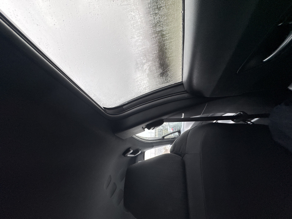 The image depicts the interior of a vehicle, viewed from a rear passenger seat looking forward towards the front passenger side and out the window, in Meudon, France. The vehicle's dark headliner is visible at the top, along with a dark fabric front passenger seat in the foreground. A black seatbelt system, including the strap and buckle, is prominent between the seat and the passenger door. The dark interior door panel features a silver-colored door handle. The side window is heavily covered in raindrops, indicating active precipitation. Through the rain-streaked window, an overcast outdoor environment is visible, with blurred forms of trees or foliage in the middle distance. Faint outlines of another vehicle are discernible through a forward-facing window beyond the front passenger seat. The overall scene suggests a rainy, overcast day.