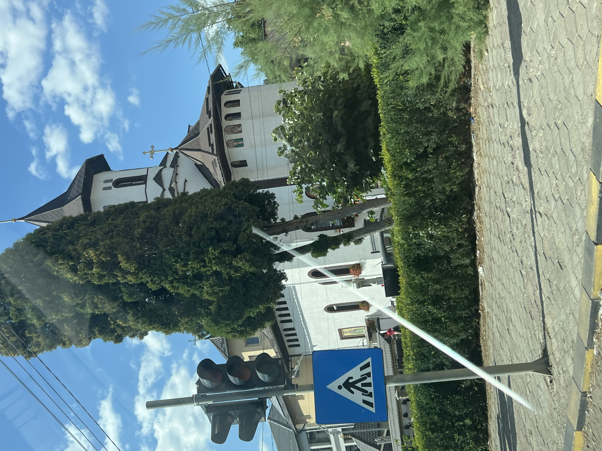 The image depicts a street scene in Gura Humorului, Romania, featuring a traffic light and a pedestrian crossing sign in the foreground. Behind these, a white church with a dark, multi-tiered roof and a cross on its steeple is visible. The church has arched windows and decorative elements above some of them. Adjacent to the church, a tall, slender green tree stands prominently, partially obscuring the building. The area is lined with a well-maintained green hedge. The ground in front of the hedge is paved with interlocking grey stones, bordered by yellow and grey curbs. The sky above is blue with scattered white clouds. In the background, other buildings and trees can be observed. The image appears to have been taken from a vehicle, as evidenced by a faint, diagonal light reflection across the scene.