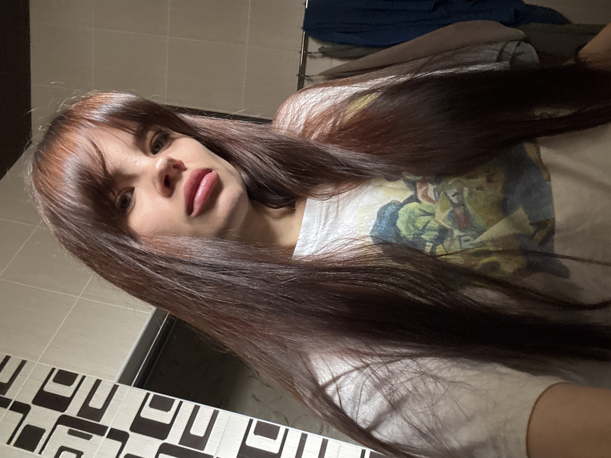 The image shows a woman with long, brown hair and bangs, wearing a white t-shirt with a colorful design. She is facing the camera with her lips slightly parted. The background includes tiled walls, possibly in a bathroom. The location in Rostov-on-Don, Russia is not confirmed in the provided image.