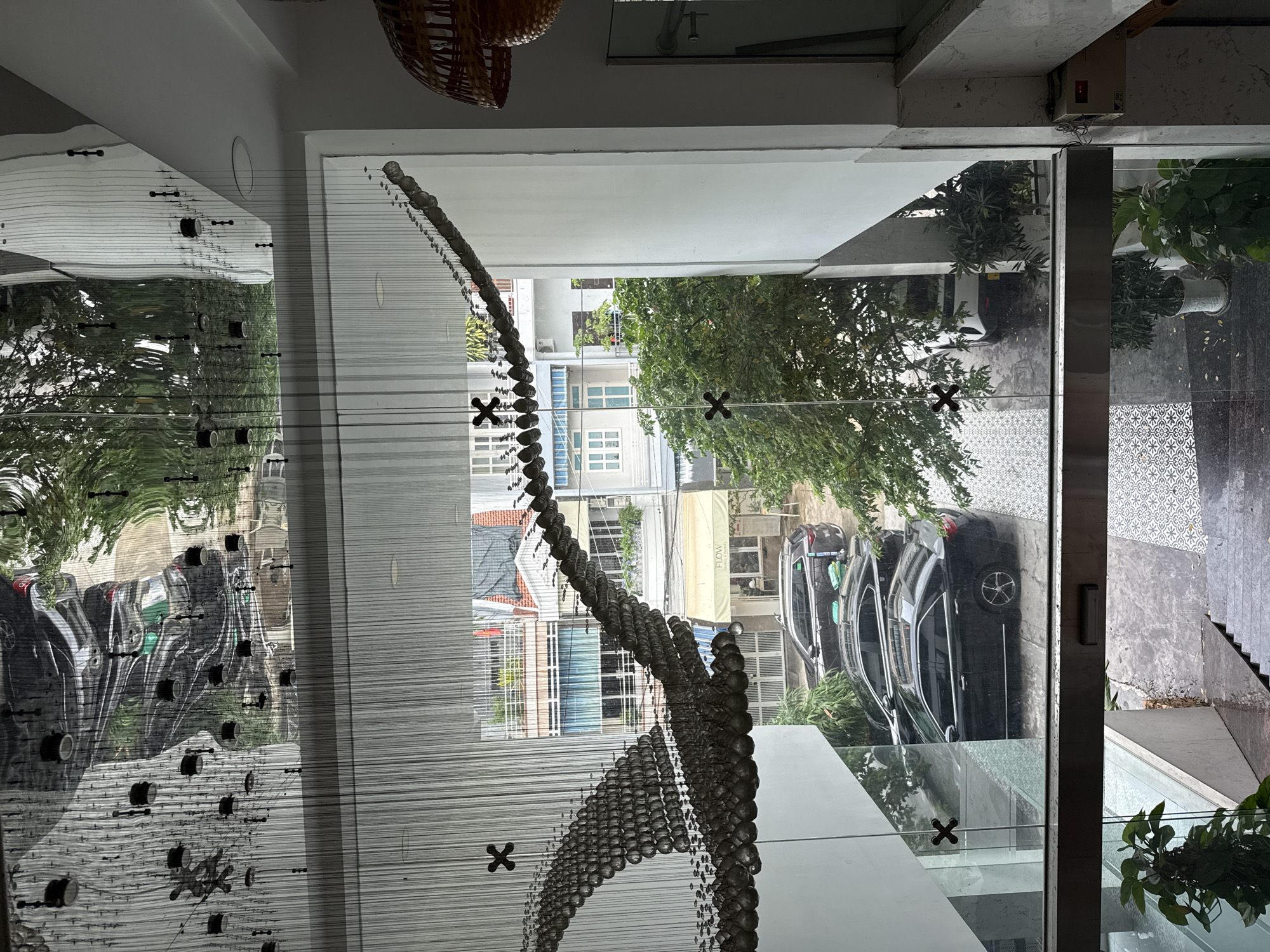 The image, taken in Bac Nha Trang Ward, Vietnam, shows an interior view through a window. The scene outside depicts a street with several parked black cars, trees, and buildings with white facades. Reflected in the interior glass is a beaded curtain or art installation and a partial view of the room's ceiling and a basket-like fixture. The window frame itself is visible, and the ground outside is concrete with patterned tiles.