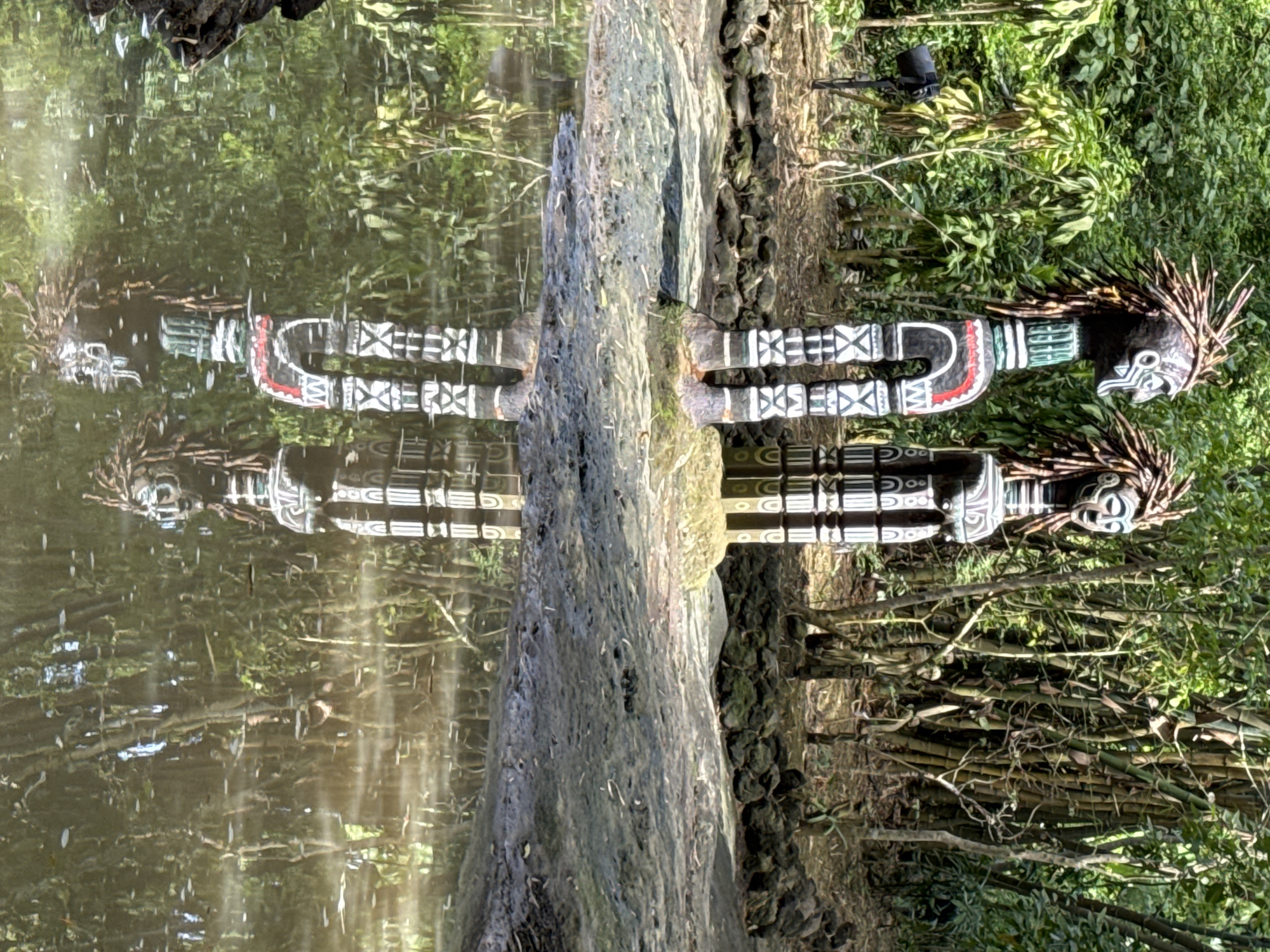 The image depicts two prominent, tall, stylized wooden statues with intricate carvings and headdresses resembling straw or twigs, situated outdoors. The statues are adorned with black, white, and red geometric patterns. They stand on a muddy, rocky bank adjacent to a body of water, where their reflections are visible. The background features dense green foliage, including trees and bamboo stalks, suggesting a tropical or subtropical environment. A black outdoor light fixture is visible to the left of the statues. There are no people or animals visible in the immediate foreground or midground. The overall scene appears to be a cultural or decorative display in a natural setting. This image is not located in the city of Kediri, Indonesia.