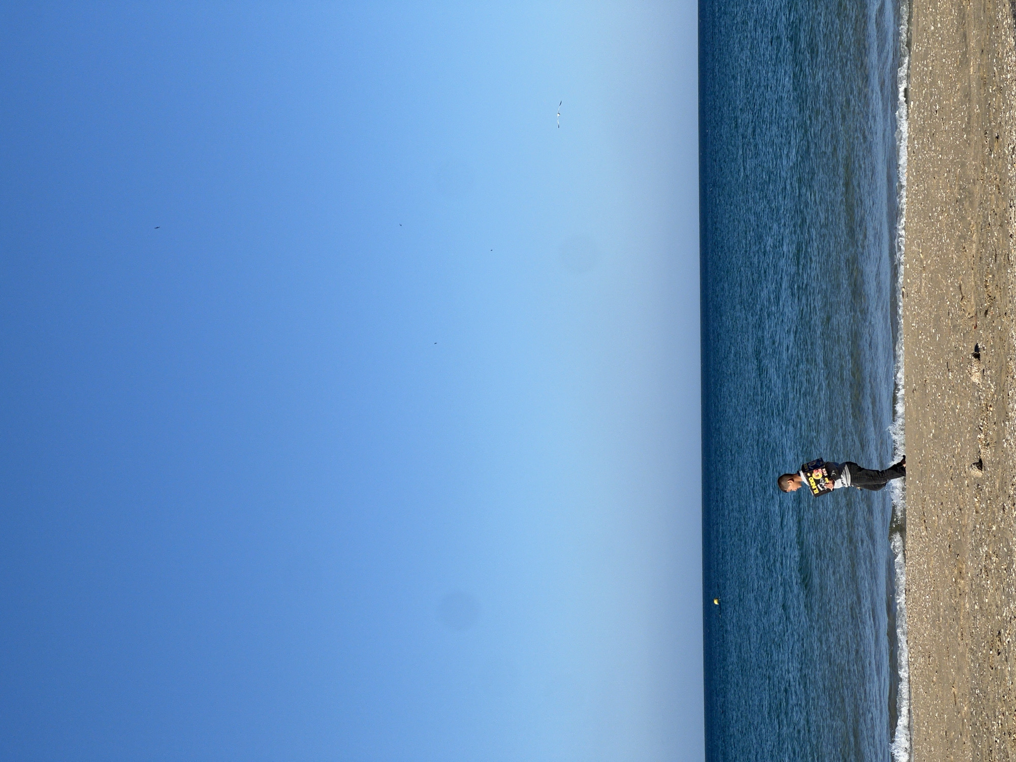The image shows a coastal scene, likely a beach in M'Diq, Morocco. The sky is clear and blue. The sea is a deep blue. A person is standing at the edge of the water on the sandy beach. The person appears to be looking at something in their hands. There are birds flying in the sky. A yellow ball is visible in the sea.