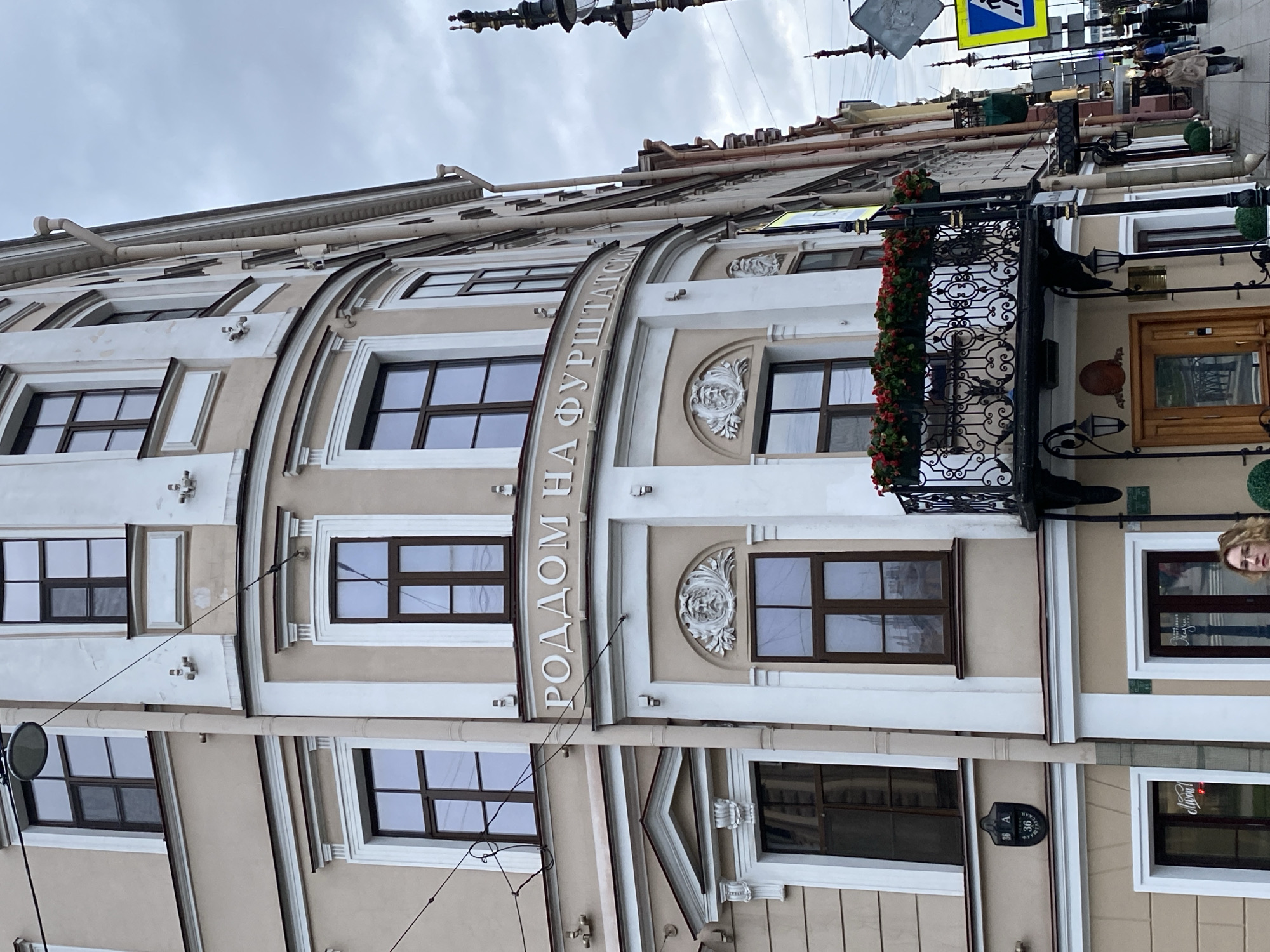 The image shows the exterior of a building in Saint Petersburg, Russia, under a cloudy sky. The building has a beige facade with white architectural details and multiple windows. Some windows are rectangular and some are arched. Along the building's side is text that reads "РОДДОМ НА ФУРШТАТСКОЙ" in vertically arranged Cyrillic letters, indicating a maternity hospital on Furshtatskaya Street. There is a decorative wrought-iron balcony with flowers, and the building appears to have historical or traditional architectural features. A traffic sign is visible at the top, along with street lights and wires. Part of a person's face is visible on the right side of the frame.