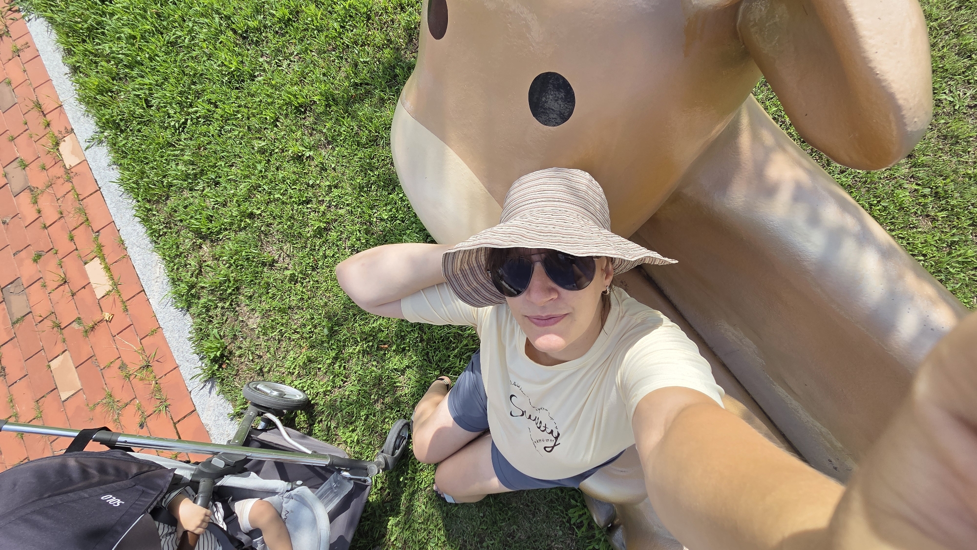A woman wearing a sun hat and sunglasses is taking a selfie while kneeling next to a large, light brown, cartoonish sculpture in a grassy area. A stroller is visible to her left, with the wheel of the stroller in the foreground. To the left of the grass is a red brick walkway with a grey border. The woman has one arm raised and resting on the sculpture, and she is looking at the camera. The sculpture appears to be a figure with two large, dark circular eyes. The ground is covered in green grass. The lighting suggests it is a sunny day. The scene is likely outdoors in a public space. The image was taken in Dunnae-myeon, South Korea.