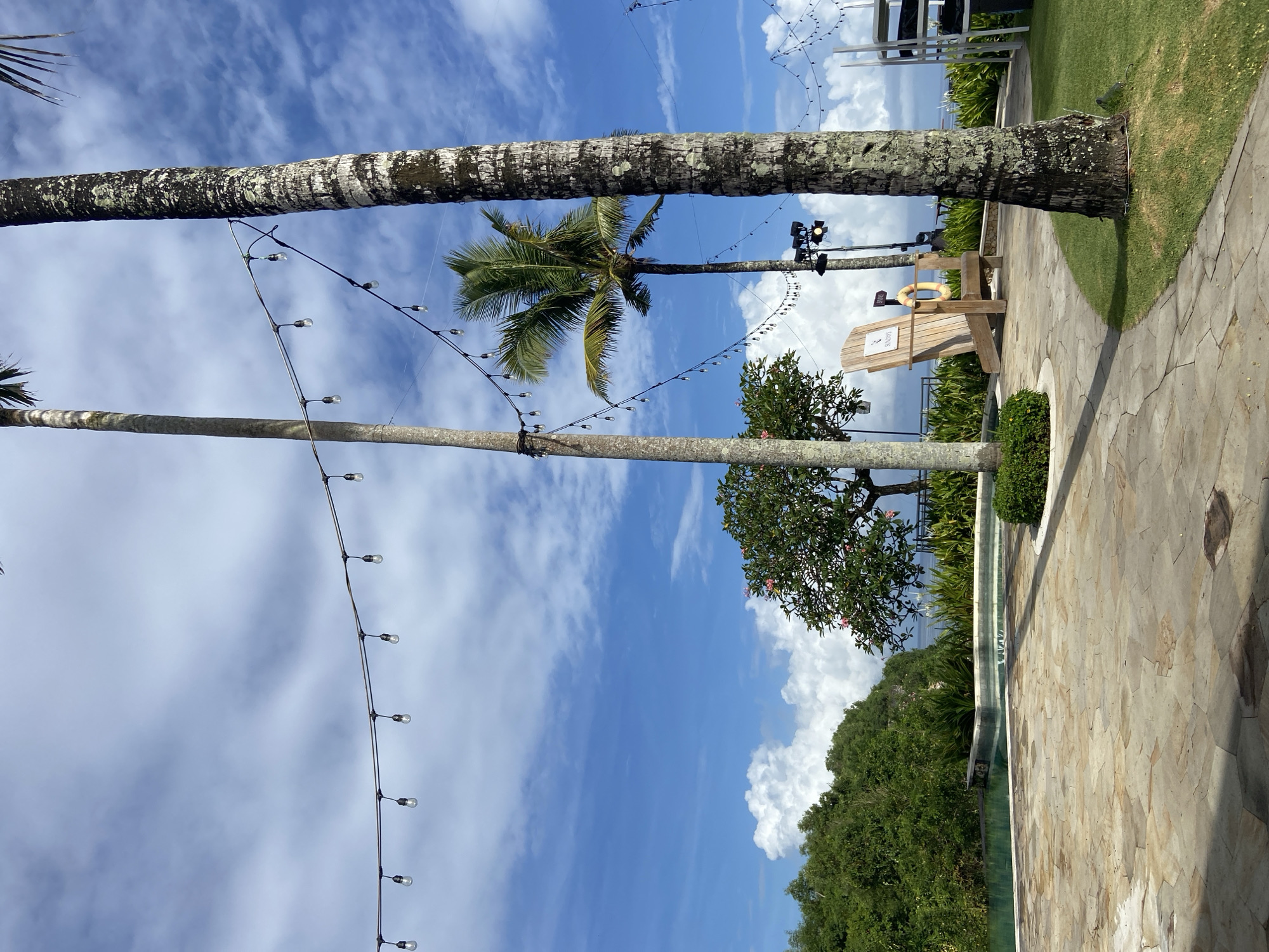 The image depicts an outdoor scene in Kuta Selatan, Indonesia, characterized by tropical vegetation and a blue sky with scattered clouds. Two tall palm trees are prominent, with string lights hanging between them. In the background, there is a wooden structure, possibly a lifeguard chair, with a lifebuoy hanging on it. Lush green foliage and trees are visible in the midground, suggesting a tropical environment. A paved pathway is in the foreground. The overall impression is a serene, tropical resort setting.