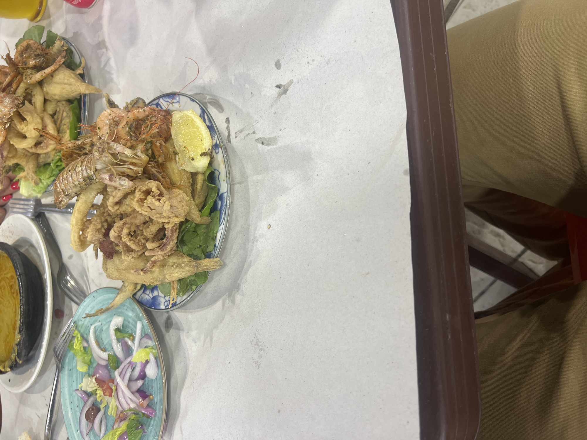 The image depicts a table in a restaurant, potentially located in M'Diq, Morocco, given the seafood dishes. The table is covered with white paper. Visible are three plates: one contains fried seafood including shrimp and calamari with a lemon wedge, another has a similar fried seafood dish, and a third contains a salad with red onions, lettuce, and tomatoes. Also, a small pot filled with a yellow substance is visible. A person wearing khaki pants sits beside the table, with the edge of the table near them. Forks are visible on the table near the plates.