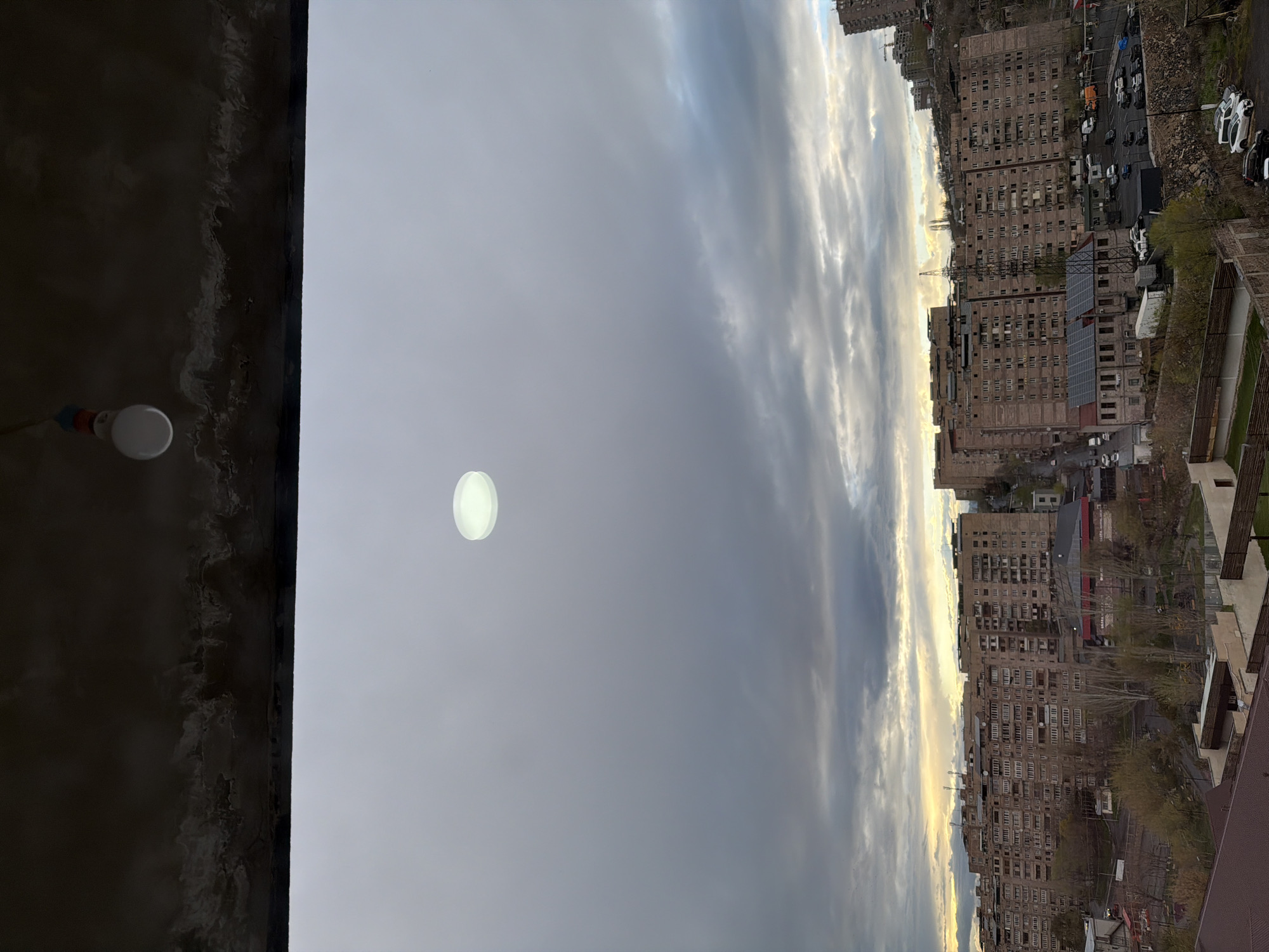 This image, taken in Yerevan, Armenia, captures an urban landscape under a cloudy sky, likely during either early morning or late afternoon given the golden light breaking through the clouds. The view appears to be from a building or balcony, with a dark, textured wall or ceiling and a white light bulb visible in the lower foreground.
The mid-ground is dominated by an expansive sky, mostly covered with grey clouds, but with clear patches towards the right where sunlight illuminates the cloud edges with warm, golden hues. A distinct circular, glowing white object, possibly a lens flare or an unidentified aerial phenomenon, is visible in the upper-left quadrant of the sky.
In the background, a dense city environment unfolds, characterized by numerous multi-story apartment buildings typical of post-Soviet architecture. These buildings are predominantly light brown or beige, featuring many windows. Streets run between the structures, and numerous cars are parked along the curbs and in open lots. Scattered trees, some still bare and others showing early signs of foliage, add a touch of green to the urban sprawl. A large array of solar panels is visible on the roof of one of the buildings, indicating a modern energy initiative. A construction crane can be seen in the distant cityscape. There are no people visible, and no discernible text.