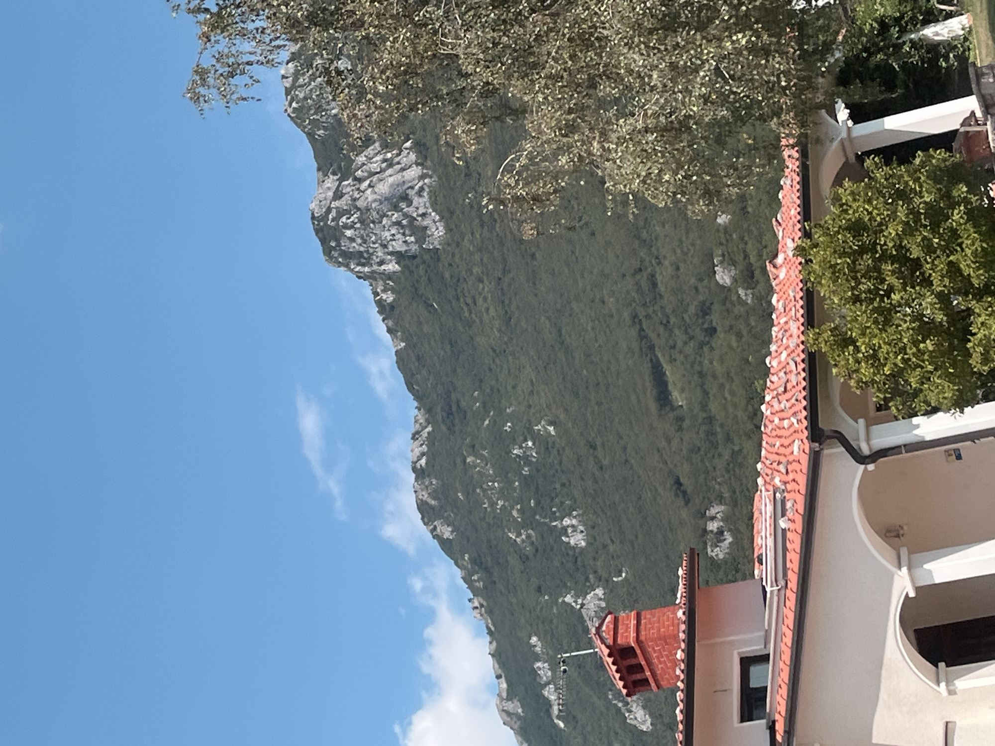 The image shows a view of the village of Budanje, Slovenia, with a house in the foreground and a mountain in the background. The house has a red-tiled roof and a red brick chimney. There is a tree to the right of the house, and the mountain is covered in greenery with some rocky outcrops. The sky is blue with a few scattered clouds.