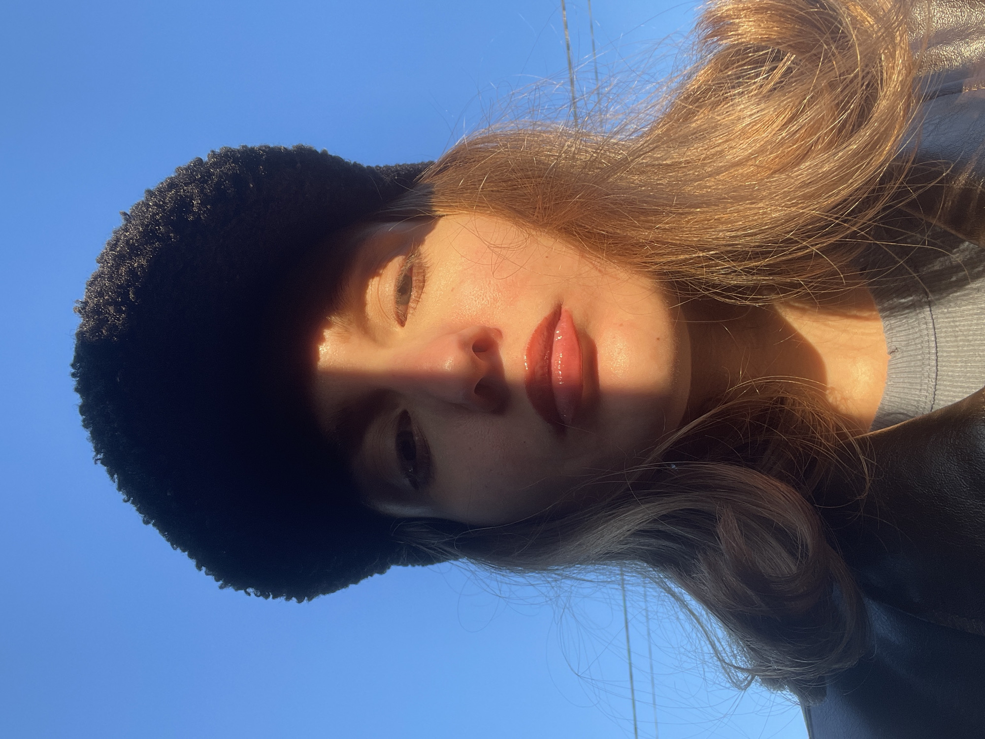 A vibrant, sunlit close-up features a young woman with her head tilted to the right, looking upwards towards a clear blue sky. She is wearing a dark, possibly fuzzy or textured, hat on the left side of her head and a dark jacket. Her light brown hair, with warm auburn undertones, is brightly illuminated by direct sunlight, appearing to flow around her head and shoulders.
A distinct, sharp shadow from the hat and her own head falls across the left side of her face, obscuring her left eye and forehead, while intensely highlighting her right eye, nose, and glossy, reddish lips. Her skin has a warm glow where touched by the sun, contrasting with the shadowed areas. Her expression is calm and neutral.
The background is an expansive, unblemished blue sky, indicating an outdoor setting on a bright, sunny day. The strong, directional light creating the dramatic shadow and warm glow suggests either early morning or late afternoon sunlight. No text is visible in the image. The scene is set in Подшивалово, Russia.