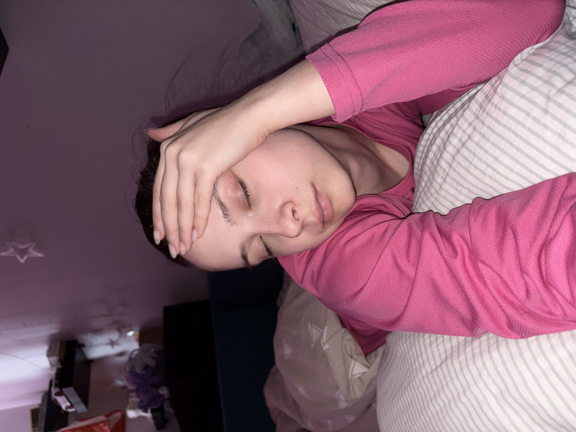 A person is lying in bed, possibly in a private residence. They are wearing a pink long-sleeved shirt. Their hand is on their forehead, and their eyes are closed. They are lying on a pillow with gray and white stripes. The room appears to be dimly lit. In the background, there is a wall, possibly with star-shaped decorative lights. The image is rotated and appears to be taken from an elevated angle. The location of the image being taken is Прикамский, Russia.