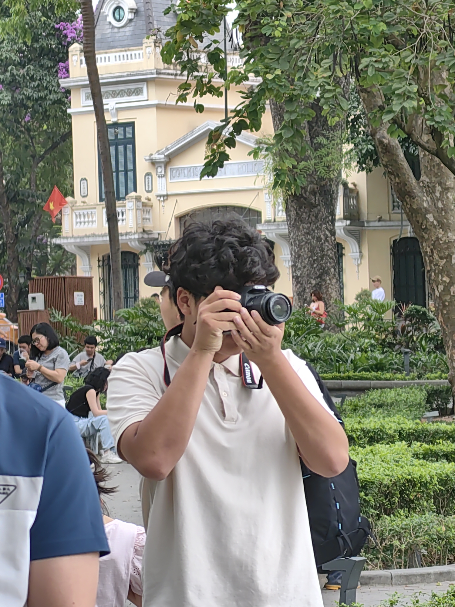 The image captures an outdoor scene in Hà Nội, Vietnam, on what appears to be a clear day. In the immediate foreground, a person, likely male and wearing a light-colored polo shirt, is focused on taking a photograph. They hold a black Canon camera with both hands, bringing it up to eye level, and a red and white "Canon" strap is visible around their neck. A black backpack is slung over their shoulder.
The setting is a public park or square, characterized by lush green foliage, including trees with mature trunks and dense bushes forming hedges. In the mid-ground, several other people are visible; some are sitting on benches, others are standing or walking, appearing relaxed and engaged in casual activities.
Dominating the background is a prominent, multi-story building of yellow and white stucco with a dark grey sloped roof. Its architecture suggests a French colonial influence, featuring ornate railings, shuttered windows, and decorative elements. A red Vietnamese flag with a yellow star flies prominently from the building's facade. Partially visible text on the building reads "COMMISSARIAT POLICE," identifying it as a police station or headquarters, consistent with the style of the Hanoi Police Headquarters. The scene conveys a calm, daytime atmosphere in a historical part of the city.