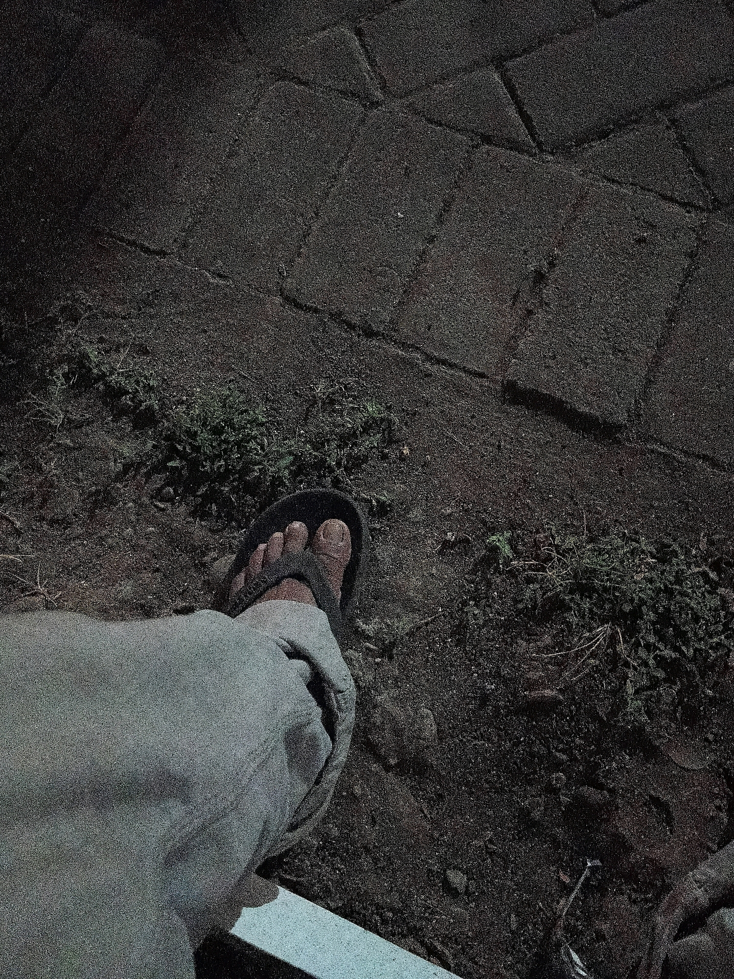 An overhead shot shows a person's right foot wearing a black flip-flop, positioned on a pathway of dark, rectangular paving stones. To the left of the foot, light-colored pants are visible. The ground surrounding the paving stones is a mix of dark soil and sparse green vegetation. The scene appears to be outdoors and is dimly lit, suggesting it might be nighttime or a heavily shaded area. The image was captured in Cikal, Indonesia.