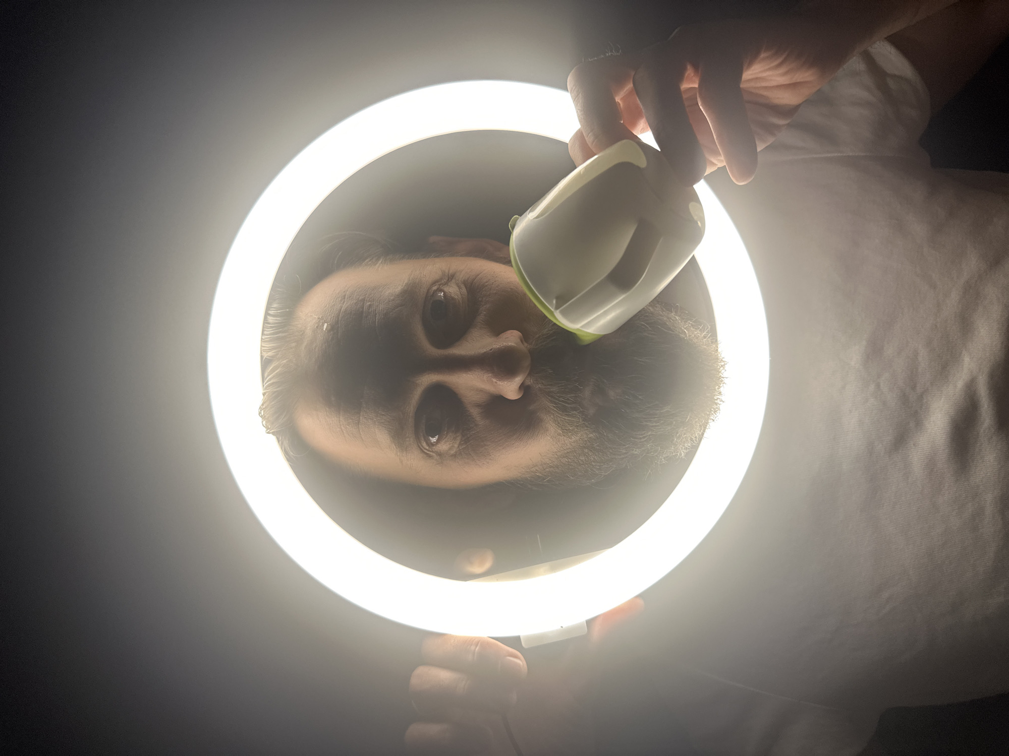 A man is holding a white and light green object up to his face within a bright circular light fixture. The light illuminates his face, which has a beard and short hair. The background is dark, except for the ring of light. The man is wearing a white t-shirt. The image is oriented so that the man appears upside down. The location is impossible to determine as there are no discernible indicators of a specific place within Fethiye, Turkey, or anywhere else.