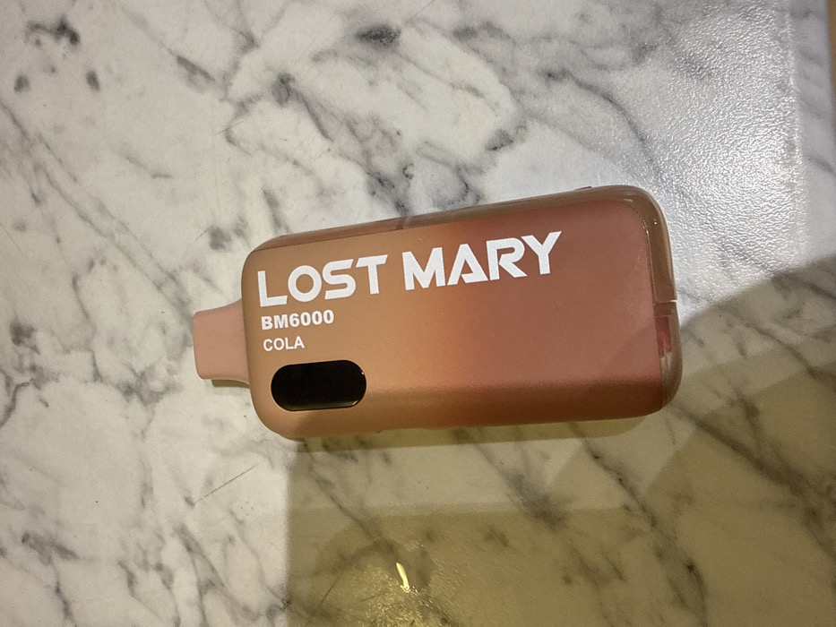 The image displays a single Lost Mary BM6000 Cola disposable vape device, positioned in Grays, United Kingdom. The device is angled on a light-colored surface characterized by a grey and white marble pattern. The vape unit features an ombre design, transitioning from a light peach tone at the integrated mouthpiece and upper section to a darker reddish-brown hue at its base. Prominent white text "LOST MARY" is displayed along the side of the device, with "BM6000 COLA" printed vertically near the top. An opaque black, elongated oval feature is present on the upper part of the device. A transparent lower section of the vape reveals a red internal component. No people, subjects, actions, or interactions are visible in the scene.