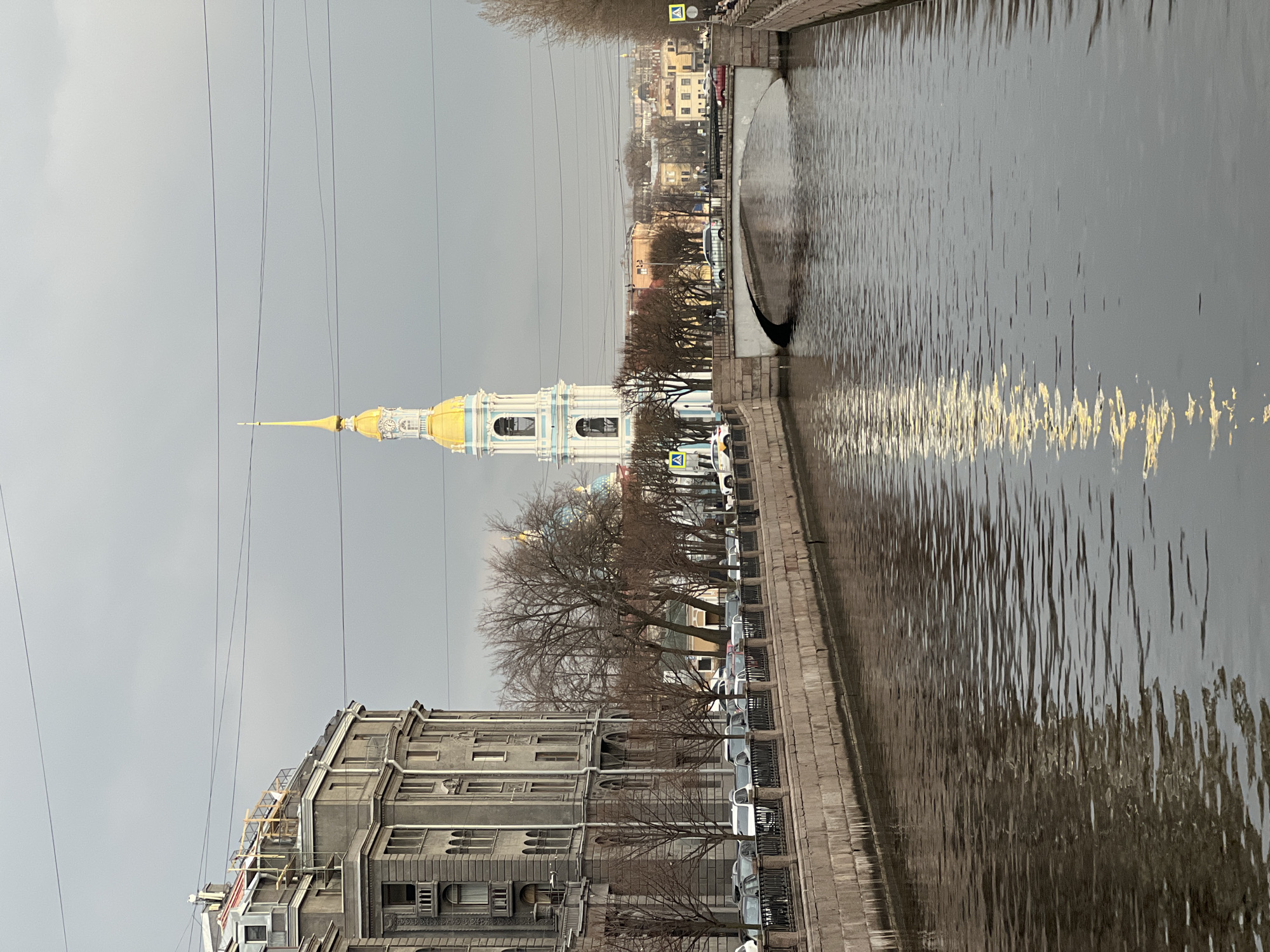 The image depicts a canal in Saint Petersburg, Russia, under an overcast sky. A prominent golden-domed cathedral with a spire is visible in the background, likely the Saint Nicholas Naval Cathedral. Buildings line the canal's edge, and bare trees suggest it's either late fall or early spring. Cars are parked along the embankment. The canal water is dark and reflects the light. A structure appears to jut into the water at the top of the frame, with water cascading in a waterfall fashion. Power lines cross the sky above the buildings.