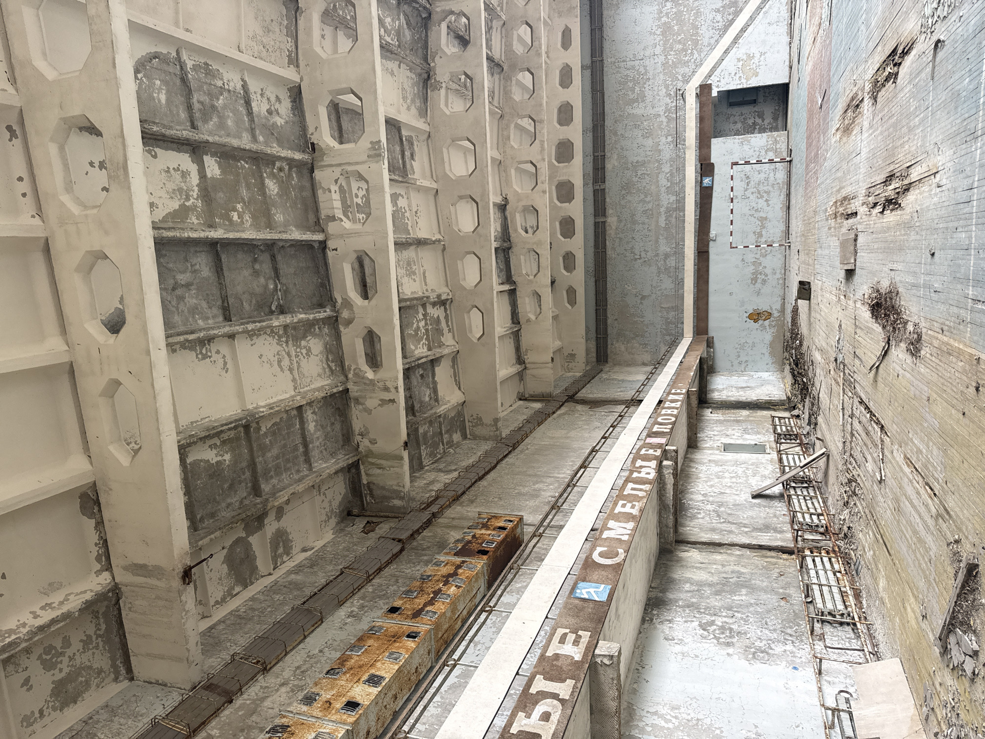 The image shows the interior of an abandoned building in the city of Prypiat, Ukraine, likely a former industrial structure. The space is characterized by its concrete construction and state of disrepair.
On the left, a series of vertical, evenly spaced concrete pillars with hollowed-out sections run the height of the image. The pillars show signs of wear, including peeling paint and exposed concrete.
The floor is cracked and marred, exhibiting layers of flaking paint. Running parallel to the left wall and extending toward the center of the image is a long, rectangular structure with Cyrillic lettering painted along its side. The lettering appears to be partially obscured. To the left of this structure is a series of rusted, open-topped boxes.
On the right, a wall shows significant decay, with sections of the brick or stone exposed and disintegrating. A series of rusted metal ladders are affixed to this wall. Further in the background, a portion of the building appears to have multiple levels or floors, with an open doorway and additional damaged walls. The entire scene conveys a sense of abandonment and urban decay.