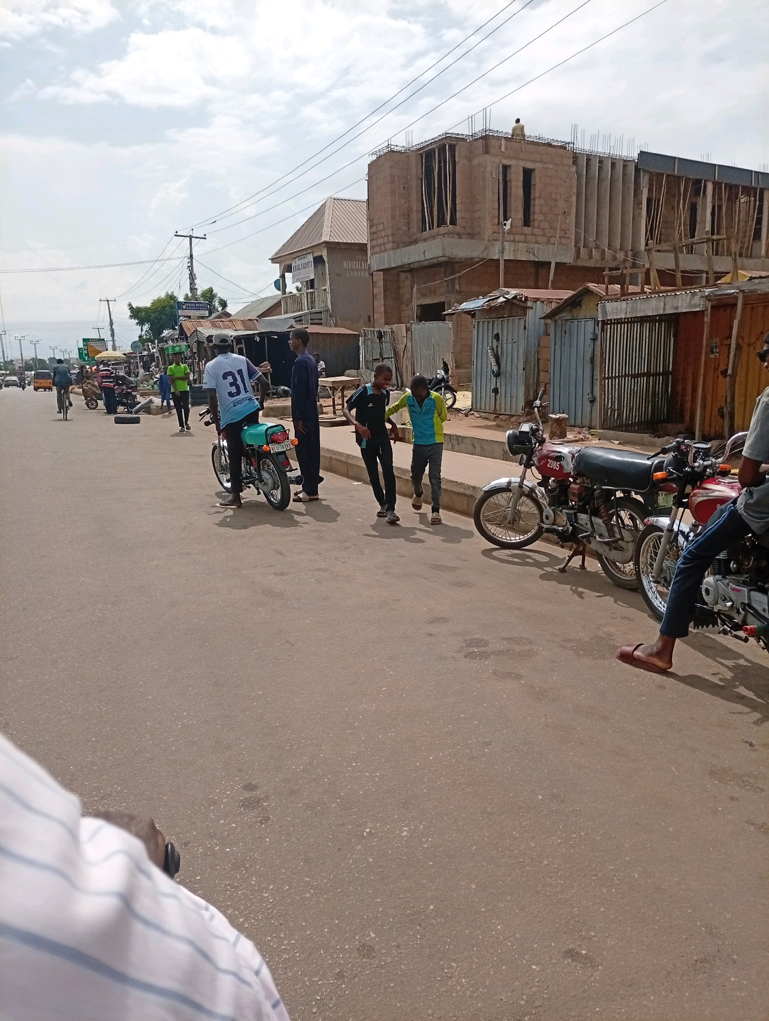 A medium shot captures a street scene in Tempure, Nigeria, under a partly cloudy sky. In the foreground, the asphalt road dominates the frame, with a white and blue striped shirt and a person's head and shoulder visible on the lower left. To the right, the lower legs and feet of a person wearing dark pants and a brown sandal are visible, casting a shadow.
Further up the street, three individuals stand near parked motorcycles. A person wearing a white t-shirt with "31" printed on the back is seated on a teal motorcycle. Next to them, a man in a dark blue robe stands. To the right, another person in a dark t-shirt and pants is seen from the waist down, leaning forward.
Beyond these individuals, a line of parked motorcycles, some with riders, stretches along the roadside. Several small market stalls with corrugated metal roofs are visible, some with various goods displayed. In the background, a building under construction, made of concrete blocks and rebar, is prominent. Traffic signs and utility poles with electrical wires are interspersed along the street. A yellow tricycle and other vehicles are seen in the distance. The overall environment depicts a bustling street scene with ongoing construction and commercial activity.