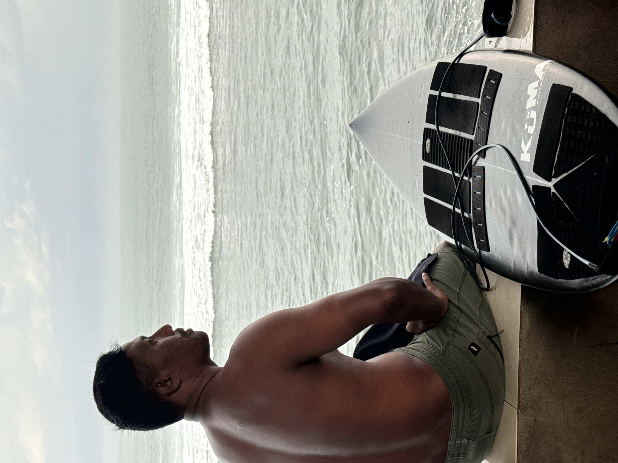 A medium, eye-level shot depicts a man seated next to a surfboard, with the ocean visible in the background. The man, who appears to be of South Asian descent, is shirtless and wearing green shorts. He is sitting upright with his head turned to the left, gazing out at the ocean. His left hand rests on his right thigh.
To the right of the man is a surfboard, which is silver and has black grip pads arranged in a pattern across its surface. The word "KUMA" is printed in white block letters on the side of the surfboard. A black leash is attached to the board.
The ocean occupies the majority of the background, with gentle waves rolling towards the shore. The sky above is overcast, creating a muted, greyish light. The scene is set on a tiled surface, possibly a patio or balcony. The location is not specified.
