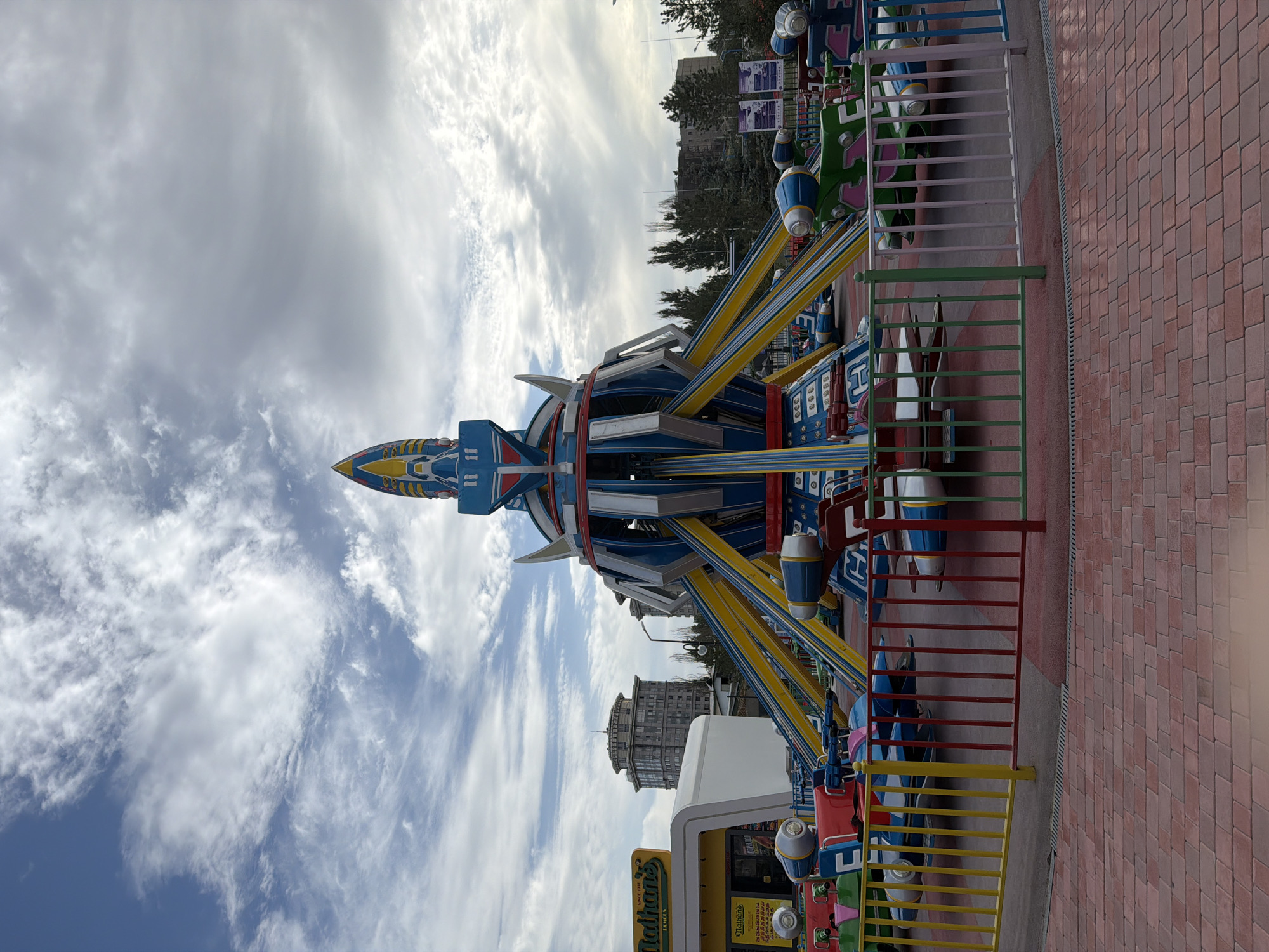 An amusement park ride with a rocket ship theme is captured from a low angle in an outdoor setting. The ride is centrally located, with multiple colorful vehicles surrounding a tall, blue, yellow, and red rocket-shaped central structure. These vehicles are designed like small spacecraft, some appearing to be red and white, and others green and pink. A multi-colored fence, featuring red, yellow, green, and blue bars, encloses the ride area. The ground in the foreground is paved with red bricks, and in the background, there are trees, buildings, and a sky filled with clouds. To the left of the ride, a yellow "Nathan's" sign is visible above a white building.