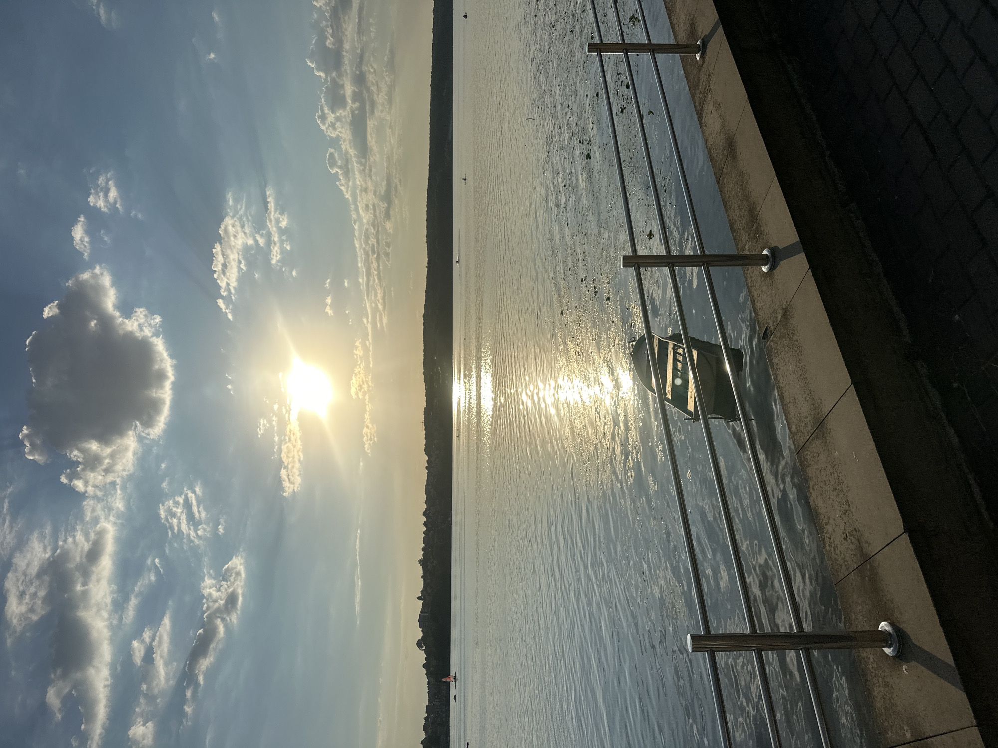 The image depicts a tranquil waterside scene at sunset in Ternopil, Ukraine. The sun, partially obscured by clouds, casts a golden glow across the calm water, creating a shimmering reflection. A lone rowboat floats near a metal railing that separates the paved walkway from the water. In the distance, the treelined shore of Ternopil lake is visible, with a few buildings nestled among the trees. The sky is a canvas of blue and white, with scattered clouds catching the last rays of the sun. The overall impression is one of peace and natural beauty.