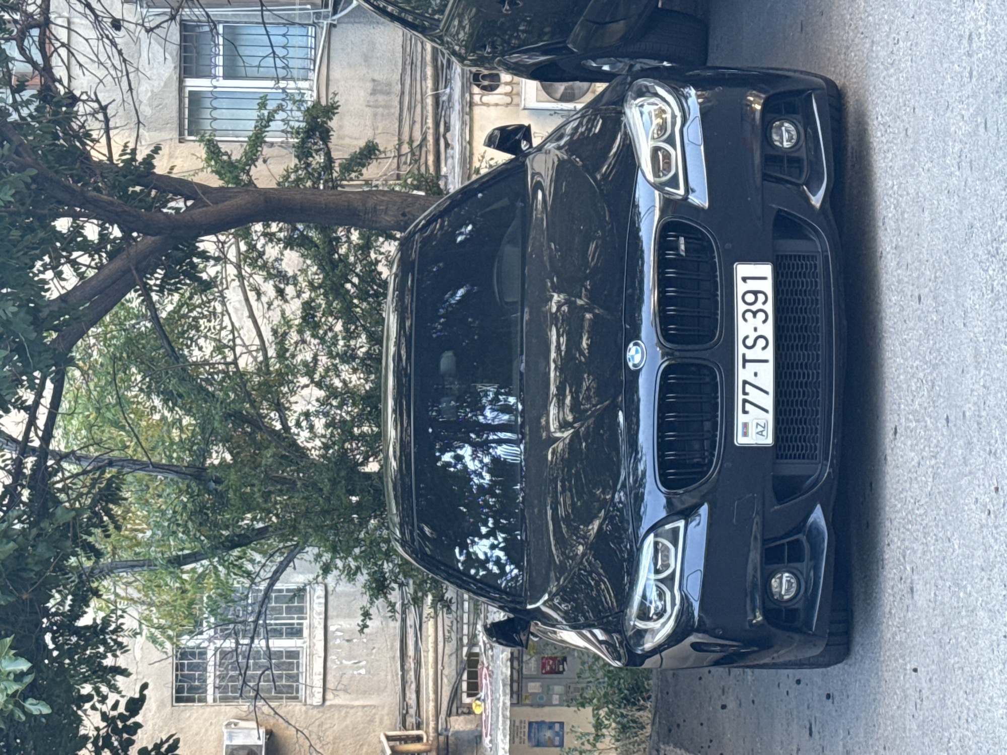 The image shows the front view of a black BMW sedan parked on a paved surface in what appears to be an urban environment. The car has its headlights on. The license plate reads "77-TS-391 AZ". In the background, there is a building with windows that have metal bars. Trees with green foliage are in front of the building, partially obscuring it. The overall impression is of a car parked on a street or in a courtyard in Baku, Azerbaijan.