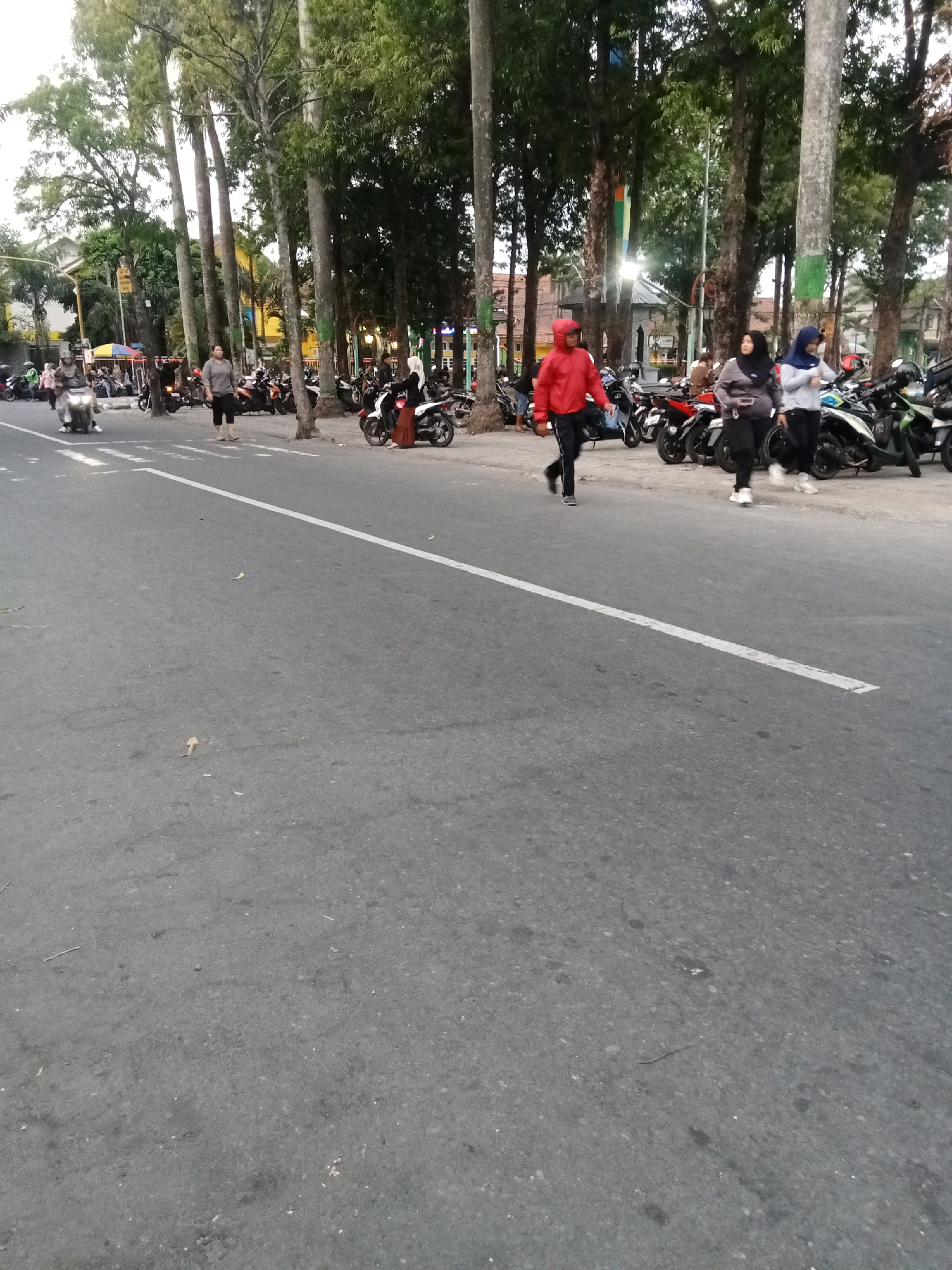 A street scene in Jombang, Indonesia, shows several people walking on the sidewalk next to a road lined with parked motorcycles. A person in a red hooded jacket and black pants walks on the road, while two women in hijabs walk on the sidewalk behind them. A white line marks the edge of the road. Tall trees with green foliage line the sidewalk. Several motorcycles are parked on the side of the road and sidewalk. In the background, buildings and more trees are visible.