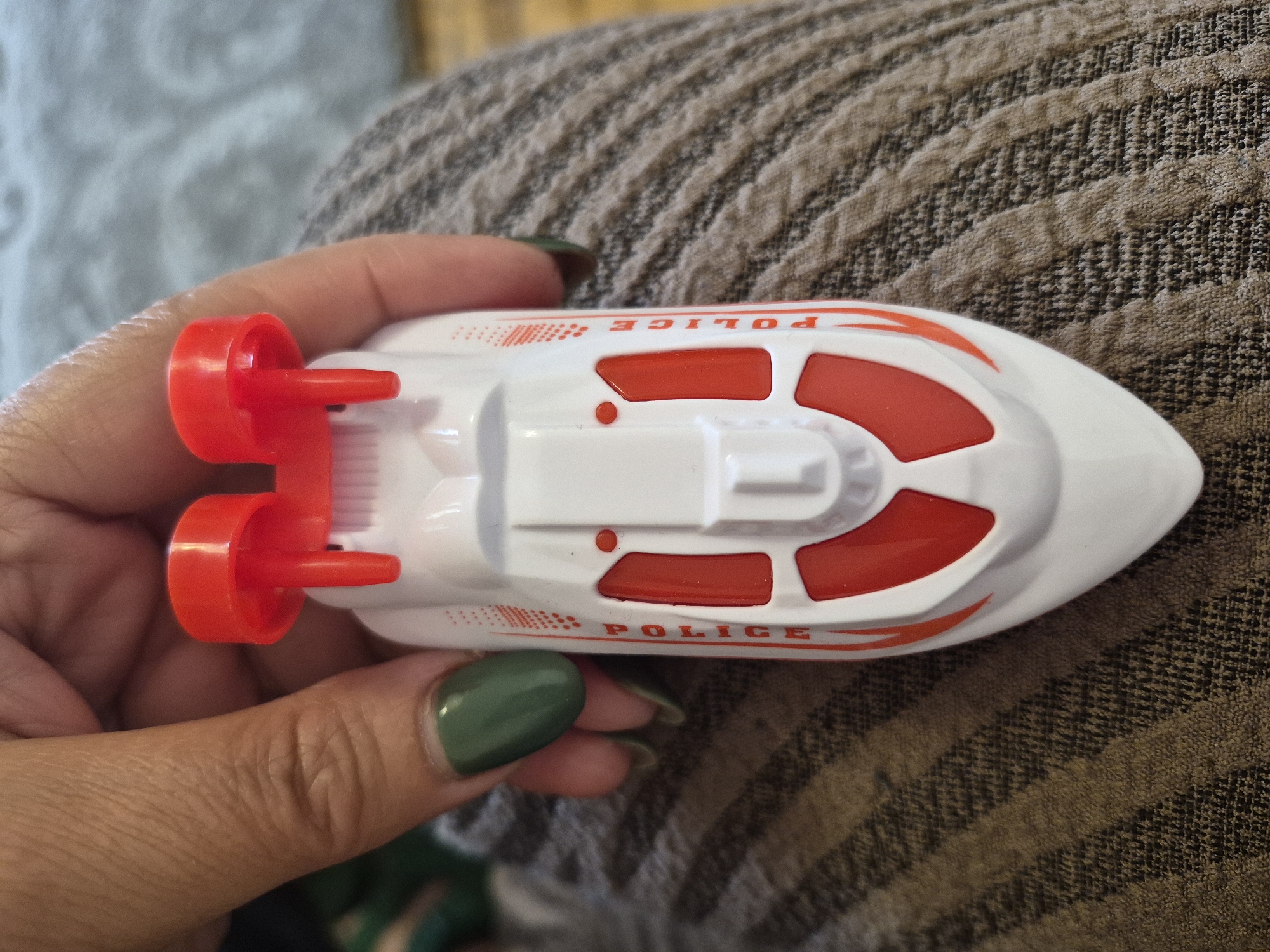 A close-up, high-angle shot captures a person's hand holding a white toy police boat. The hand, visible on the left side of the frame, has dark green nail polish on the fingernails. The toy boat is primarily white with prominent red details, including two red propellers at the back. Red-colored sections are integrated into the boat's top deck. The word "POLICE" is clearly printed in red capital letters on the side of the hull, accompanied by a red dotted pattern. The toy is held above a textured fabric surface, possibly a blanket or clothing, which features a brown and beige striped pattern. A lighter, out-of-focus fabric, likely gray, is visible in the background. The lighting suggests an indoor setting. No specific event or strong emotions are discernable. No explicit location cues are present in the image itself, though the context indicates Yerevan, Armenia.