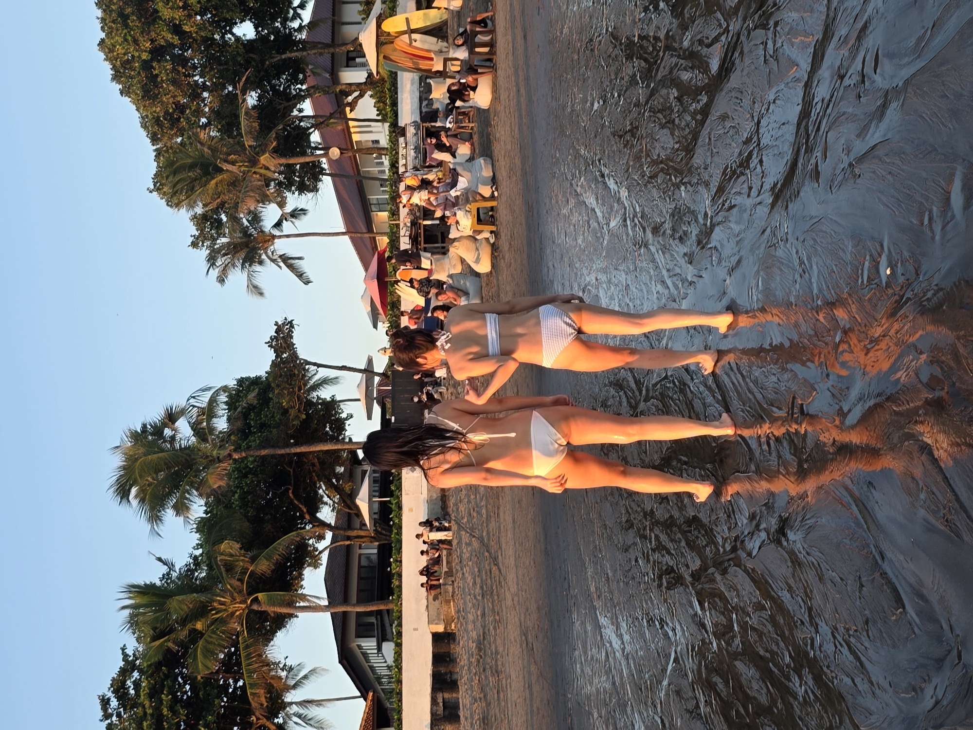 In North Kuta, Indonesia, two women in bikinis walk on a wet beach near palm trees and buildings. The woman on the left wears a white bikini, while the woman on the right wears a white and blue striped bikini. They are walking towards the ocean, with their backs to the camera. The beach is sandy and wet, with reflections of the sky and trees in the water. In the background, there are people sitting on beanbag chairs, umbrellas, surfboards, and buildings. The scene is illuminated by the warm glow of the setting sun.