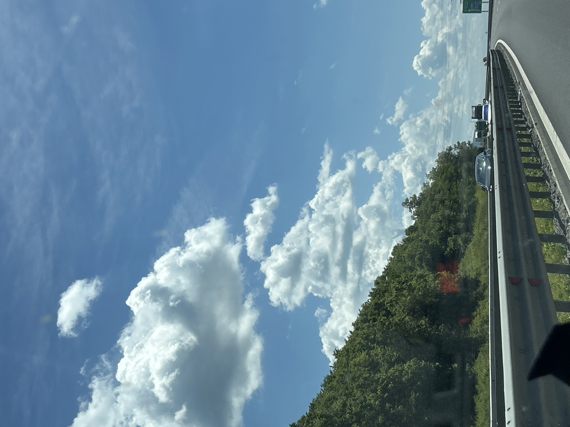 The image depicts a highway scene under a bright blue sky with fluffy white clouds. The highway curves to the right, with a metal guardrail separating the lanes. On the left side of the highway, a densely wooded hill slopes upwards. Several vehicles are visible on the highway, including cars and trucks. The lighting suggests it is daytime, likely late morning or early afternoon, given the shadows and the brightness of the sky. The environment appears to be rural or suburban, with the natural landscape dominating the roadside. There are no discernible people visible in the image. The image was taken from a vehicle moving on the highway, as indicated by the perspective and the partial view of a car's interior in the foreground. The location is specified as the city of Rajana, Bulgaria.