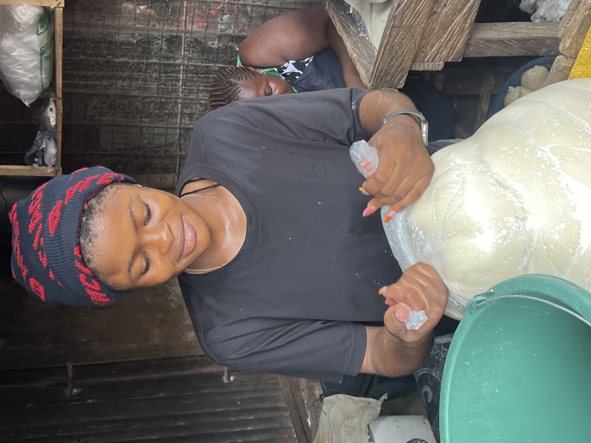In Nkpor, Nigeria, a young woman wearing a black t-shirt and a red and black patterned beanie is seen in what appears to be an open-air market stall. She is holding a large, translucent plastic bag filled with a white, dough-like substance, and her hands are partially submerged in it. Her fingernails are painted with bright colors, and she has a delicate gold chain around her neck. Her expression is one of focused engagement with the task at hand. To her right, another woman with braided hair is visible in the background, also seemingly engaged in market activities. The environment is rustic, with wooden structures and shelves holding various items, including bags of what might be ice or other goods. A large green bucket is positioned to the left of the young woman. The scene captures a moment of commerce and daily life within a Nigerian marketplace.