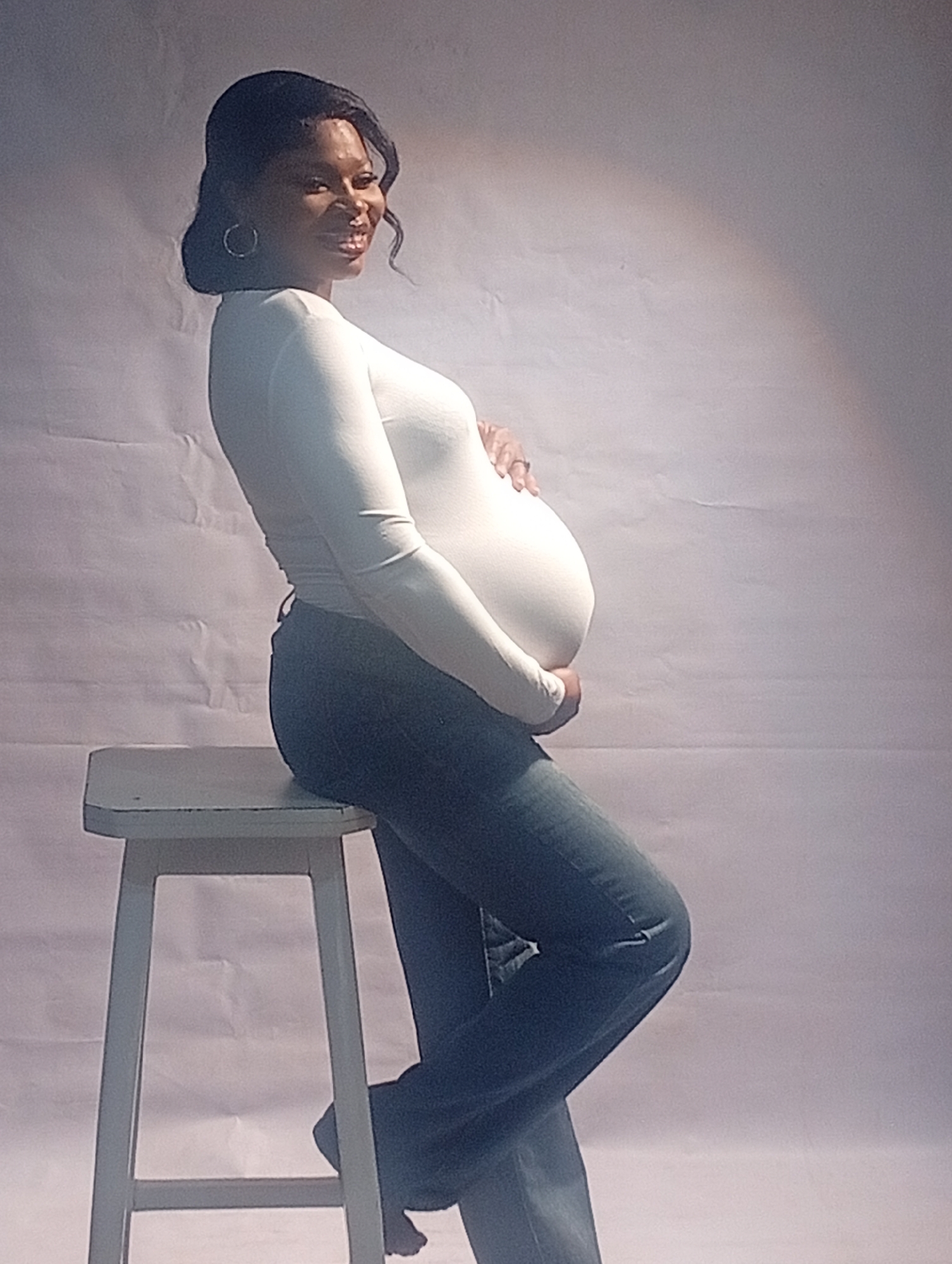 A pregnant Black woman is posing for a photo in a studio. She is wearing a long-sleeved white shirt and jeans. She is sitting on a white stool and cradling her belly. The background is a plain white backdrop. There is no visible evidence to place the location in Barakin Akawo, Nigeria.