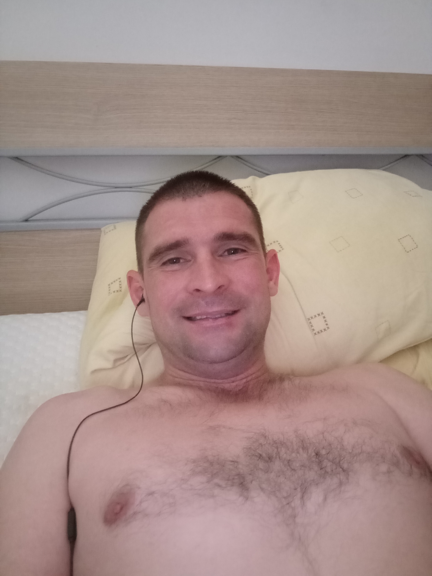 The image shows a man lying in bed, looking at the camera. He is shirtless and wearing black earbuds. The man has short brown hair and a short beard. He is smiling. Behind him is a yellow pillow with a diamond pattern. The bed is covered with a white sheet. The headboard of the bed is visible in the background. The lighting suggests it is daytime. There is no visible text in the image. The location context is Civitanova Marche, Italy, but there are no specific visual cues in the image to confirm this.
