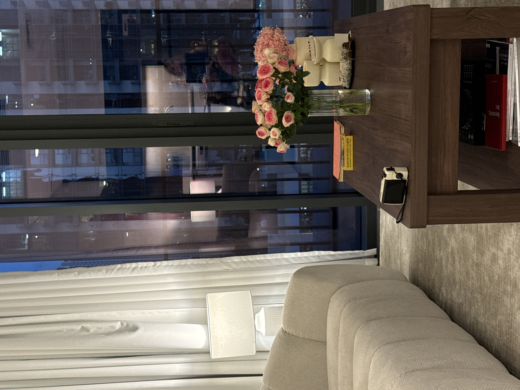 The image shows a living room scene at dusk or night, potentially in Dubai, United Arab Emirates, as suggested by the high-rise buildings visible through the large window. The window dominates the left side of the frame, reflecting the interior lighting and some details of the room itself. White, pleated curtains hang in front of part of the window.
A beige-colored, deeply tufted sofa occupies the lower portion of the frame.
A dark brown, wooden side table stands to the right. On the table, there is a bouquet of pink roses in a clear glass vase, several small white ceramic objects, and a small box of "Albano-Waite Tarot" cards. A black and silver camera sits near the edge of the table. There are books and magazines on the lower shelf of the table.
Outside the window, tall buildings with many windows are visible. Some are lit, while others are dark. The reflection in the glass shows the room's interior and a blurred figure. The lighting in the room is soft and warm, with a table lamp visible.
