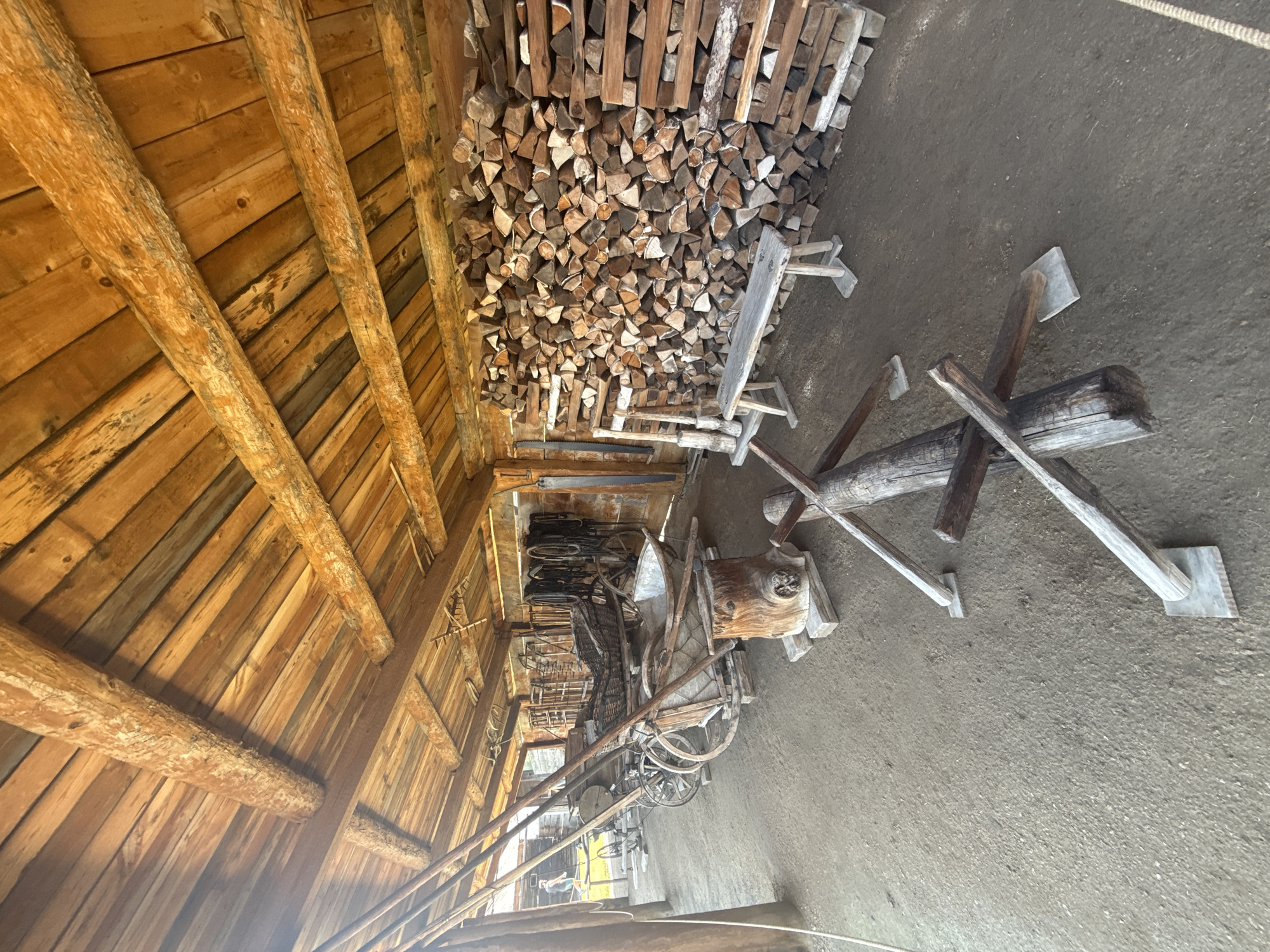 This image depicts the interior of a rustic wooden structure, likely a barn or workshop, located in Shushenskoye, Russia. The scene is filled with historical artifacts and supplies. In the foreground, a large pile of chopped firewood is stacked against the back wall. To the left, several horse-drawn carriages and carts are parked, some appearing old and weathered. A wooden bench sits in front of the firewood. Various tools and equipment, including what appear to be agricultural implements and harnesses, are visible throughout the space. The ceiling is constructed of rough-hewn wooden beams and planks, contributing to the primitive atmosphere. The floor is dirt. A person is faintly visible walking in the background through an open doorway on the left.
