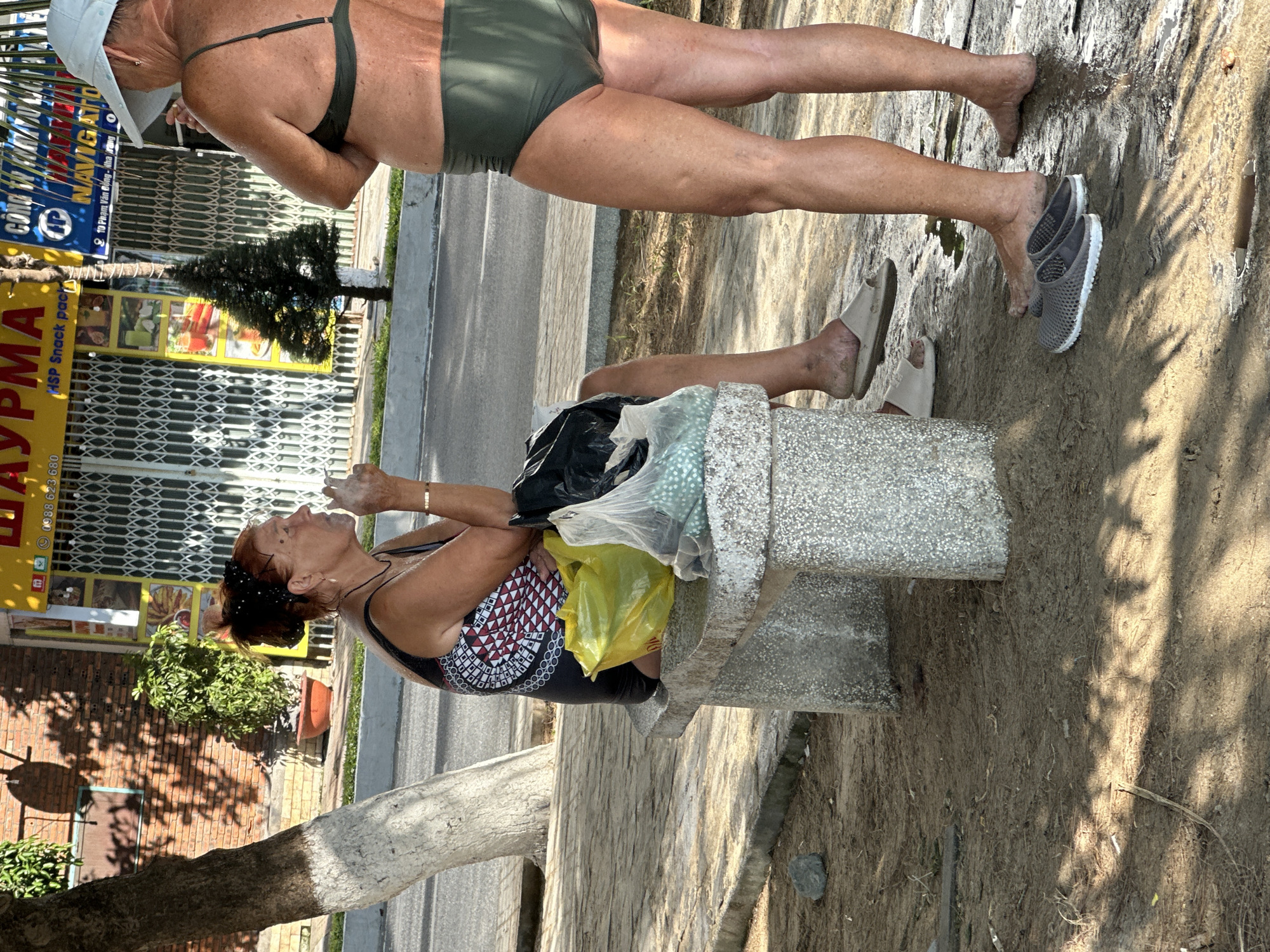 The image captures an outdoor scene, appearing to be in the Bac Nha Trang Ward, Vietnam, as indicated by the signage. Two women are featured in the frame. One woman is standing near the edge of a paved surface, possibly a street or walkway, and is wearing a two-piece olive green swimsuit and a white hat. The other woman is seated on a light-gray stone bench, wearing a black and red patterned top. She appears to be interacting with something, possibly eating, as she is looking upward with her hand near her mouth. Shopping bags are placed on the bench next to her. The surroundings include a building with a yellow sign displaying the word "УРМАТ" and a telephone number. There is also a smaller sign with the text "NAVIGATO" and "2 10 Phạm Văn Đồng - Nha Trang". A small Christmas tree is visible near the building. The area is partly shaded by a tree, and the ground appears to be covered with soil or sand.