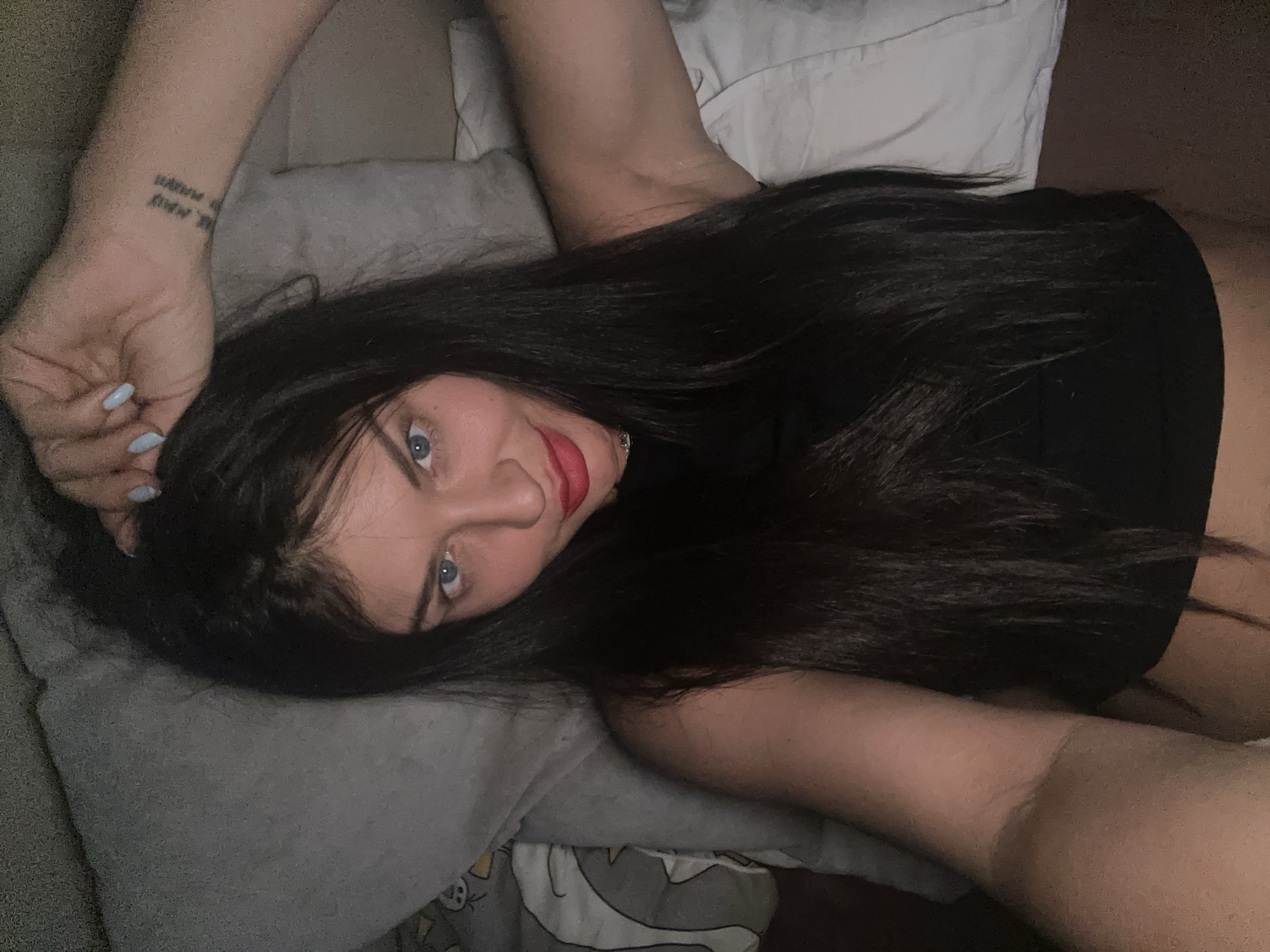 A woman with long, dark hair and blue eyes is captured in a close-up, top-down shot, lying down and looking directly at the camera with a gentle smile. Her dark hair is fanned out around her head and shoulders. She is wearing a black top and has prominent red lipstick and light blue nail polish. Her left arm is visible, bent at the elbow, with her hand resting near her head. On her left inner forearm/wrist, there is a tattoo featuring two lines of text. The first line appears to read "be. me" or "be. may", and the second line is largely obscured and difficult to decipher clearly, possibly starting with "always". She is resting on what appears to be a bed or couch, with a gray pillow under her head and another with a cartoon-like pattern partially visible in the bottom right corner. The lighting is dim and soft, suggesting an indoor evening or nighttime setting. The subject's expression conveys a relaxed and engaging mood. The provided context indicates the location is Moscow, Russia.