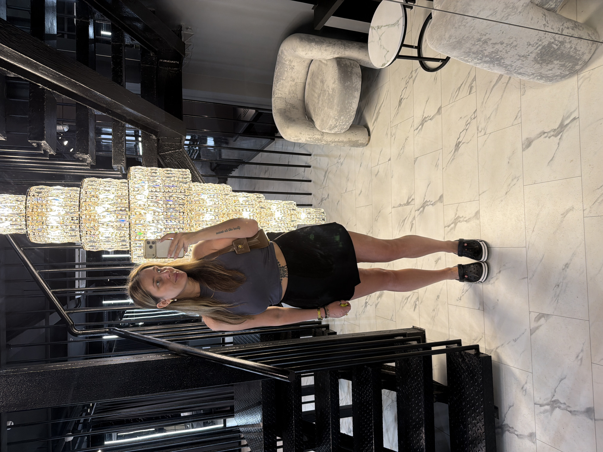 In the image, a woman stands on a marble-tiled floor, taking a selfie with her phone. She is wearing a grey cropped tank top, a black skirt, and black clogs. A brown shoulder bag is slung over her left shoulder. To her left is a black metal staircase with industrial-style steps and railings. To her right, a plush, light-colored armchair sits next to a small round table with a marble-patterned top. A large, multi-tiered crystal chandelier hangs prominently in the background, illuminating the scene. The overall aesthetic suggests an indoor, likely commercial or residential, space with a modern and luxurious design. There is no information to suggest the location is in Укромновское сельское поселение, Ukraine.