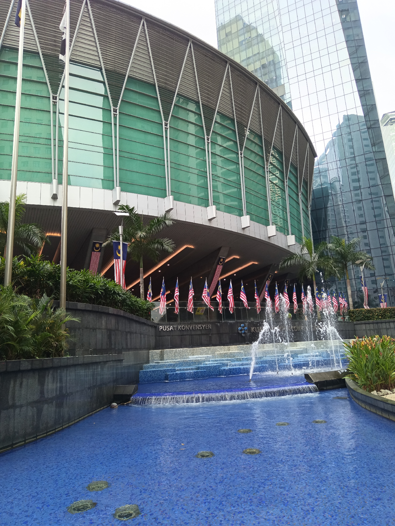 The image shows the exterior of the Kuala Lumpur Convention Centre (KLCC) in Kuala Lumpur, Malaysia. The convention centre features a large, curved glass facade with a metallic roof structure. In front of the building, a multi-tiered fountain is in operation, with water spraying upwards. A series of Malaysian flags are displayed along the facade and on flagpoles. Lush green plants and palm trees are visible, adding to the outdoor environment. In the background, modern skyscrapers with glass exteriors reflect the sky and surrounding buildings. The foreground of the image shows a large pool of water with a blue tiled surface and submerged circular features. The overall scene depicts a prominent architectural landmark in a bustling urban setting.