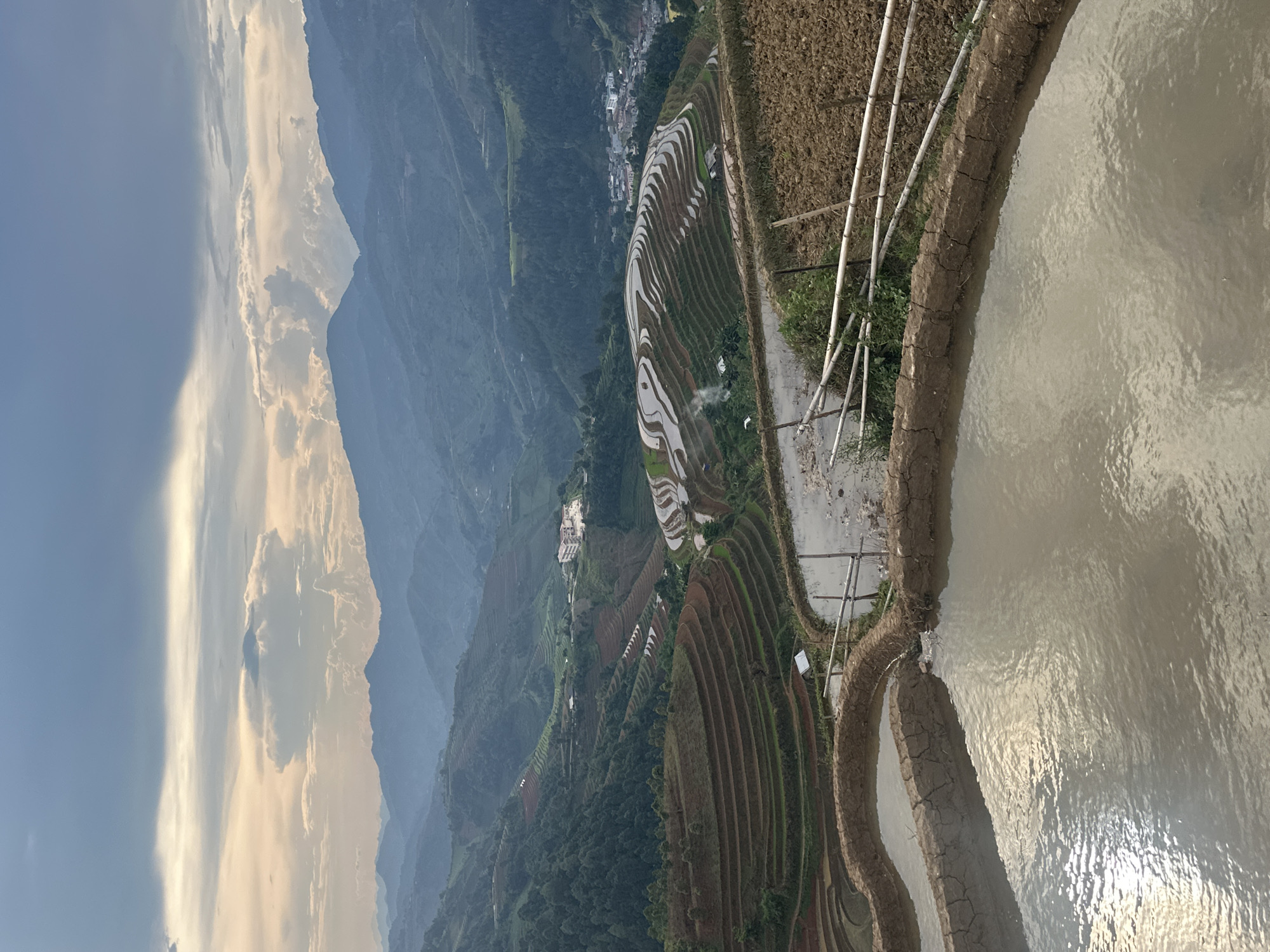 This captivating, wide-angle landscape photograph depicts the vast and intricate terraced rice fields of a mountainous region, likely in Lao Chai, Vietnam, given the provided context. The image is captured from an elevated perspective, looking across multiple layers of terraces that cascade down a steep valley.
In the foreground, a large, shallow body of water, likely a flooded rice paddy, dominates the right side. Its surface subtly ripples, reflecting the ambient light. A distinct, cracked earthen embankment separates this water from a dry, brown field on the immediate left, where several light-colored, slender poles (resembling bamboo) are loosely propped up. Small patches of green vegetation can be seen near these poles.
Beyond the foreground, the main subject of the image unfolds: numerous terraced fields. Some terraces appear dry, displaying rich, reddish-brown earth, while others are filled with water, creating mirror-like surfaces that reflect the sky and highlight their serpentine curves across the landscape. Further into the mid-ground, the terraces blend with the verdant slopes of distant mountains, which recede into a soft, atmospheric haze, suggesting significant depth. A small collection of buildings, possibly a village, is nestled amongst the terraces in the mid-distance.
The sky occupies the upper portion of the frame, showing a clear, pale blue on the left that transitions into dramatic, sunlit clouds on the right. These clouds are bright and voluminous, suggesting either late afternoon or early morning light, casting a warm glow on parts of the scene and creating strong reflections in the water-filled paddies. The overall impression is one of tranquility and the impressive scale of human endeavor harmonizing with the natural environment. No visible text is legible in the image.